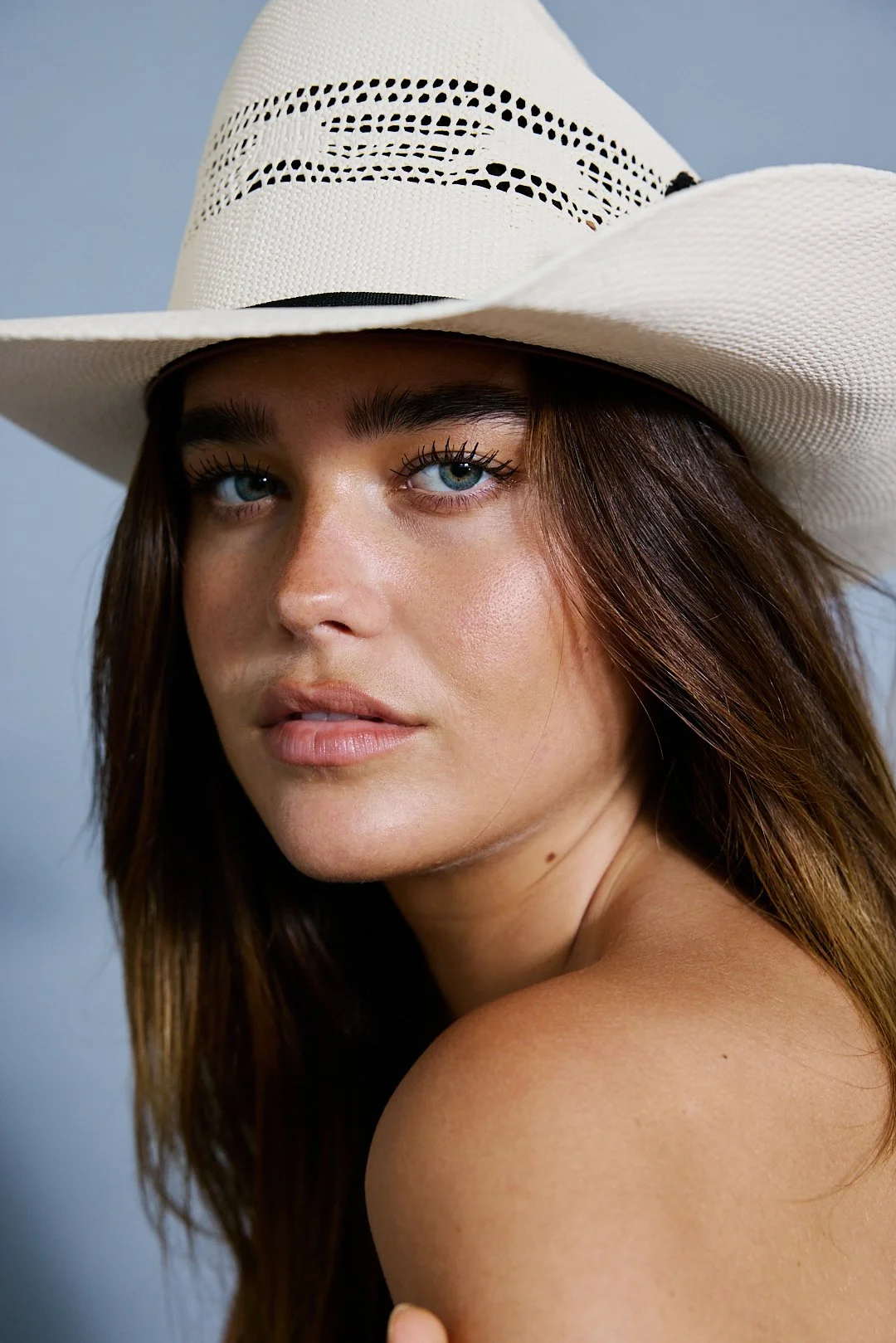 A woman with blue eyes and brown hair wearing a white wide-brimmed hat, looking at the camera with a neutral expression.