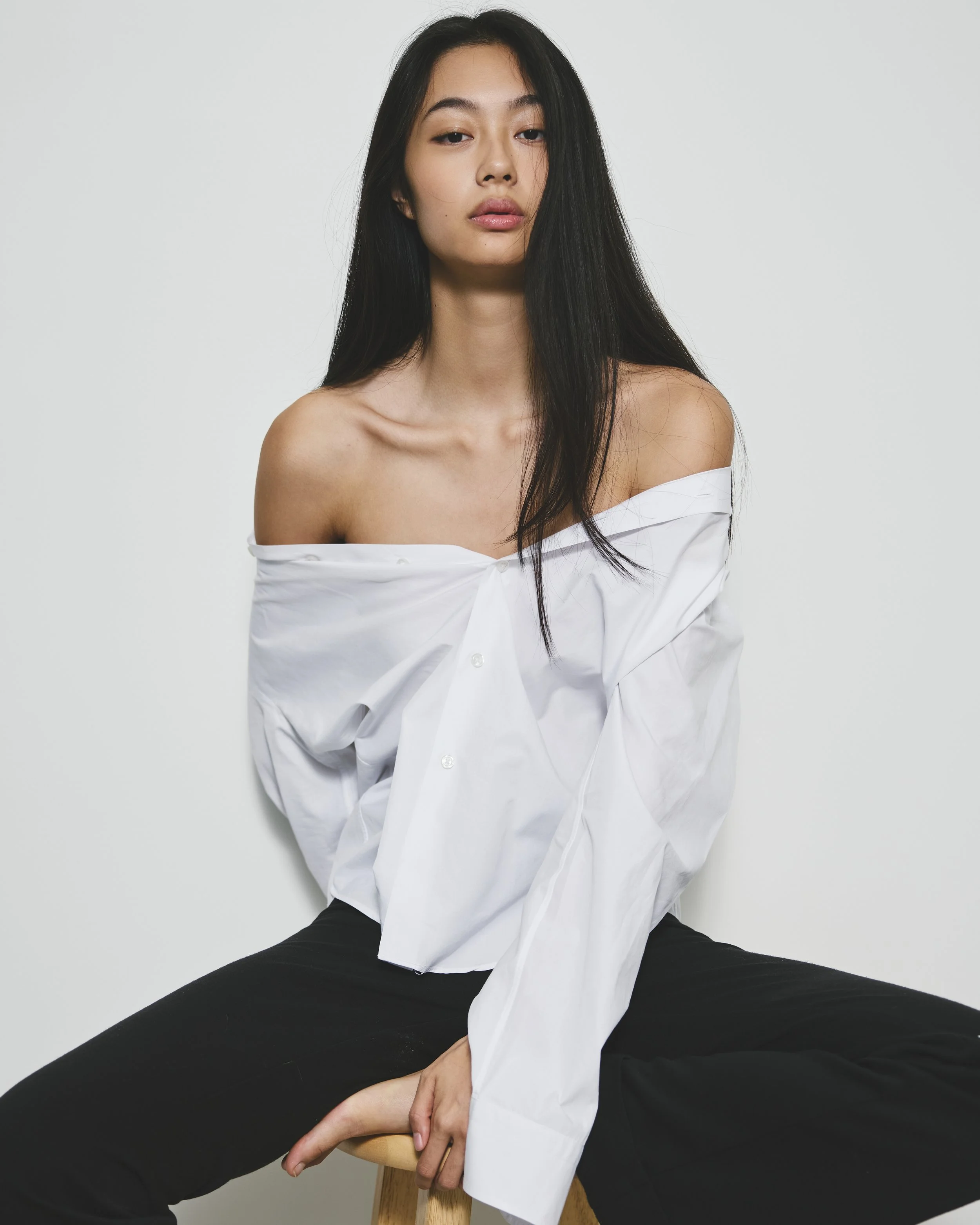 A woman with long black hair and fair skin, sitting on a wooden stool against a plain white background, wearing an off-the-shoulder white shirt and black pants.