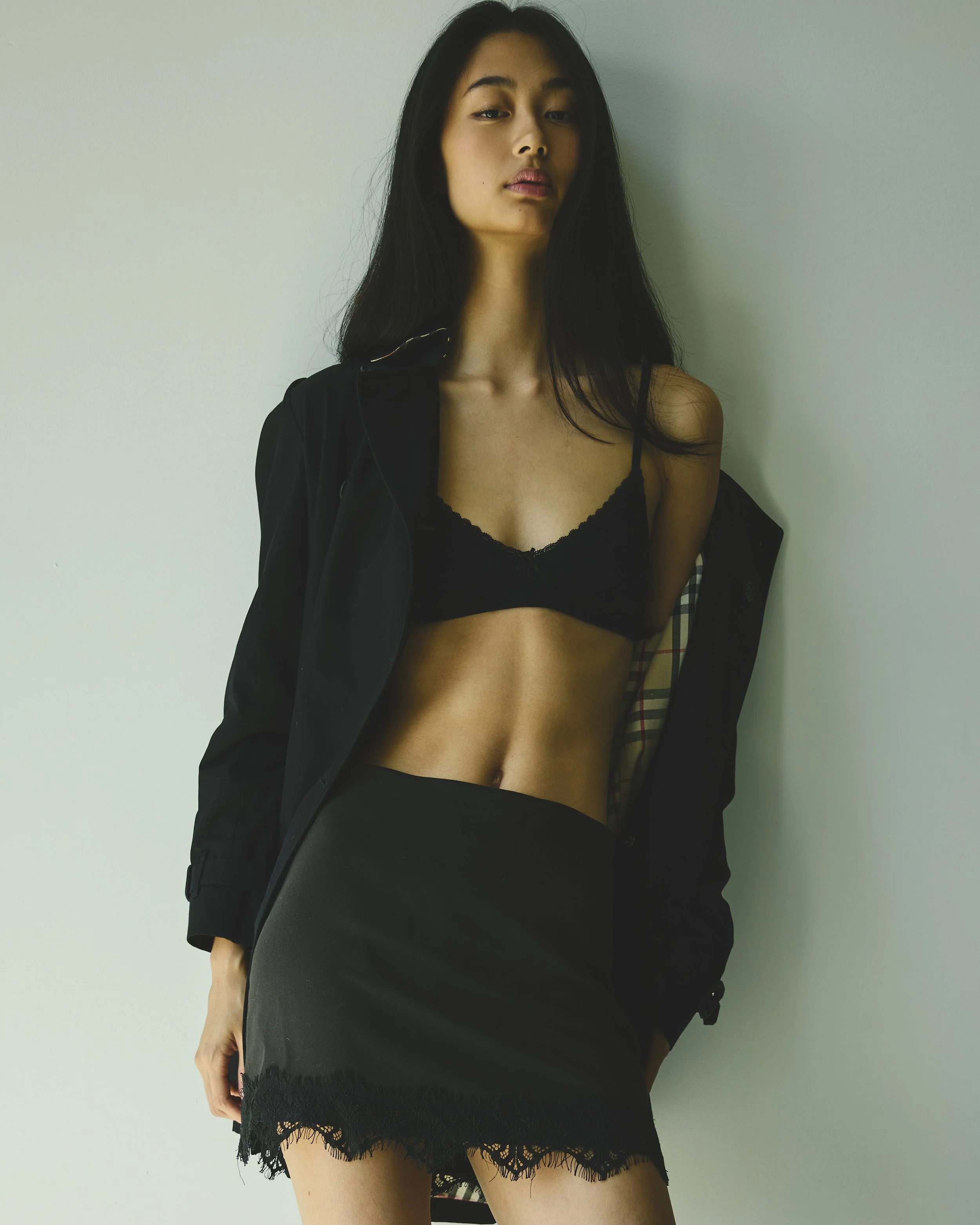 A woman with long dark hair wearing a black lace bralette, black lace skirt, and a black jacket. She is standing against a plain light-colored wall, slightly turned with her head tilted and looking into the camera.