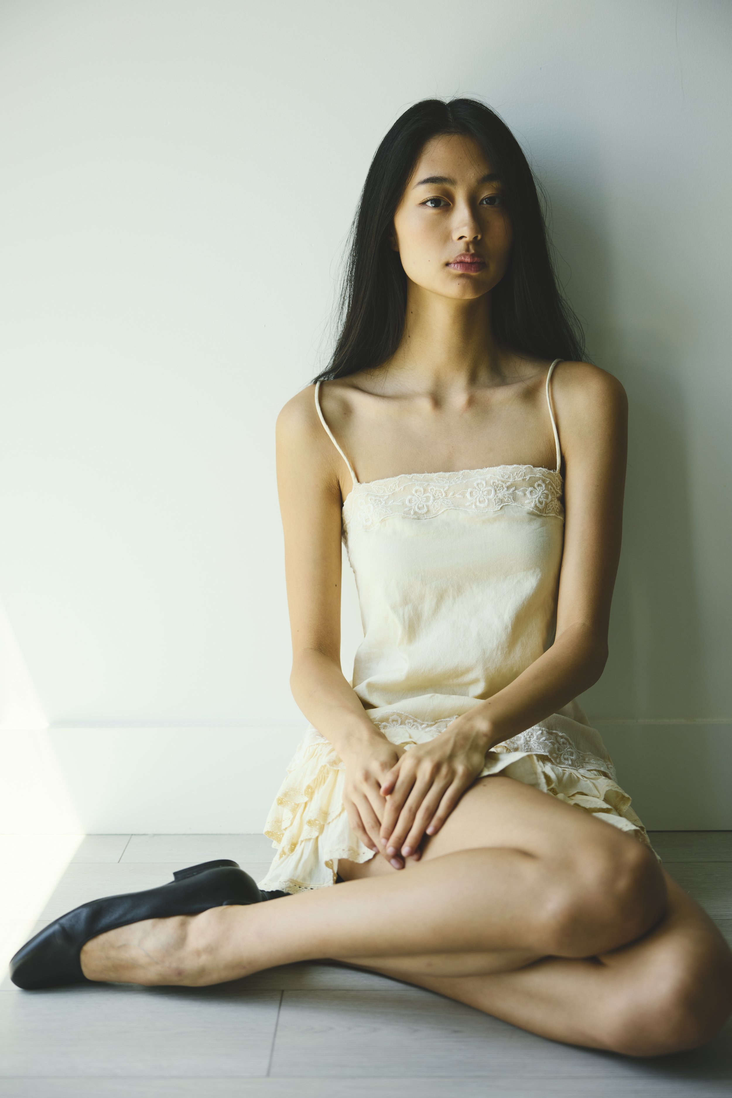 A woman sitting on the floor with her legs crossed, wearing a cream-colored dress and black shoes, in front of a plain white wall.