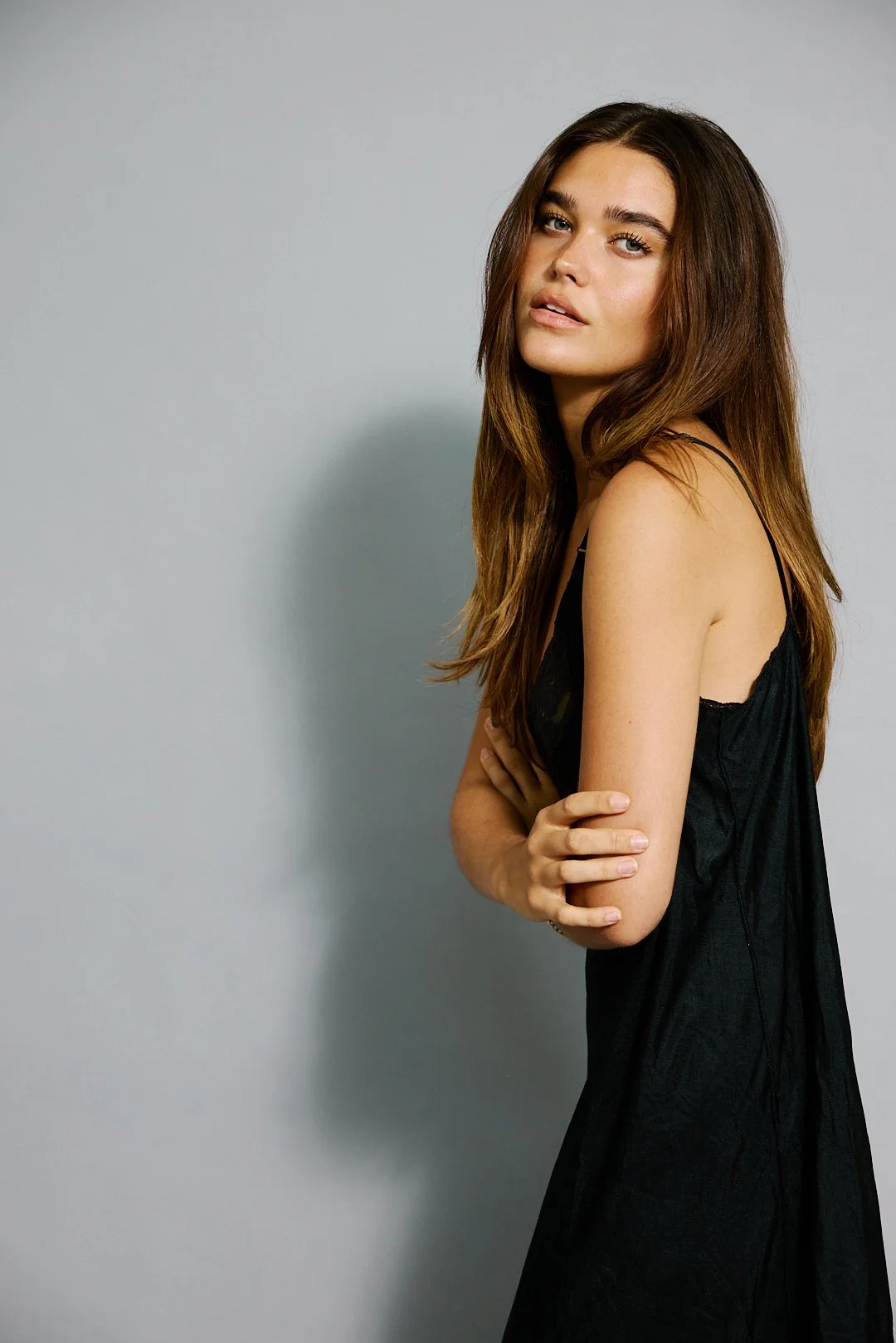 A young woman with long brown hair, wearing a black sleeveless dress, standing with her arms crossed against a plain light gray background.