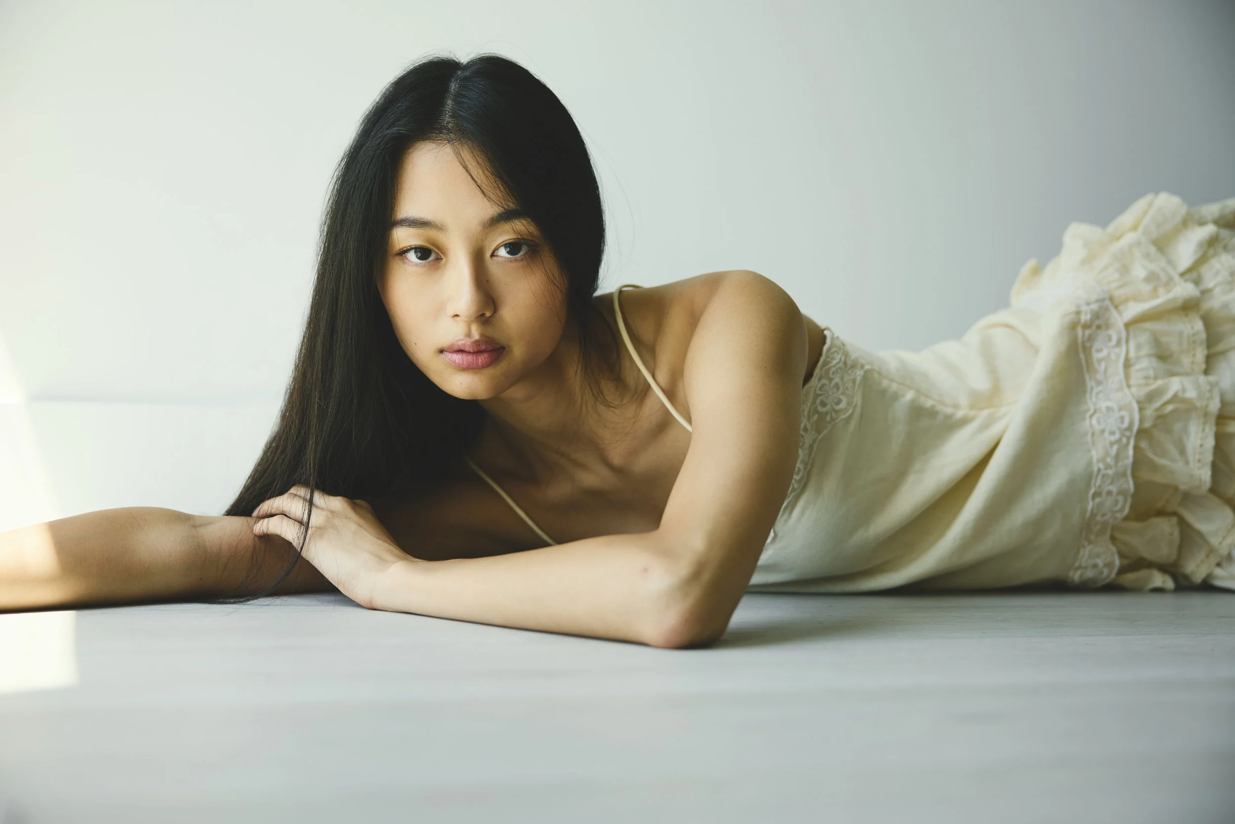 A young woman with long black hair lying on her side on a white surface, wearing a light-colored dress with lace details, in front of a plain white wall.