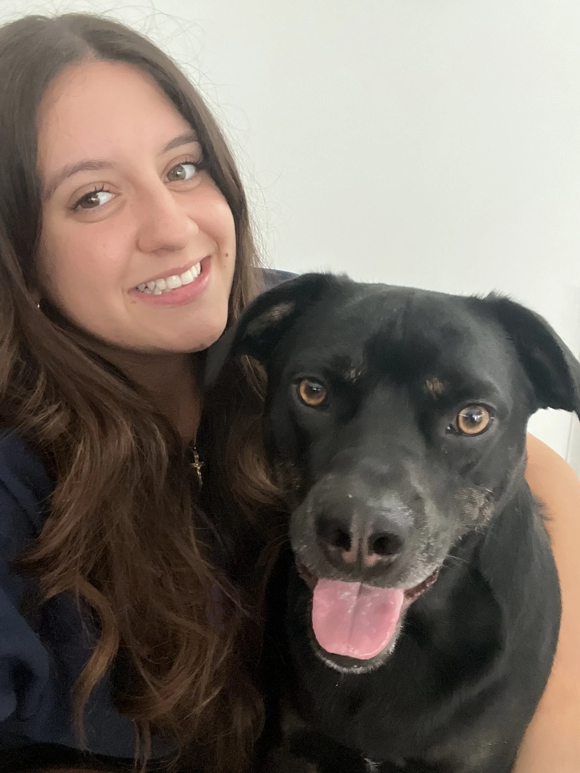 A young woman with long brown hair smiling and taking a selfie with a large black dog that has brown eyes and tongue out.