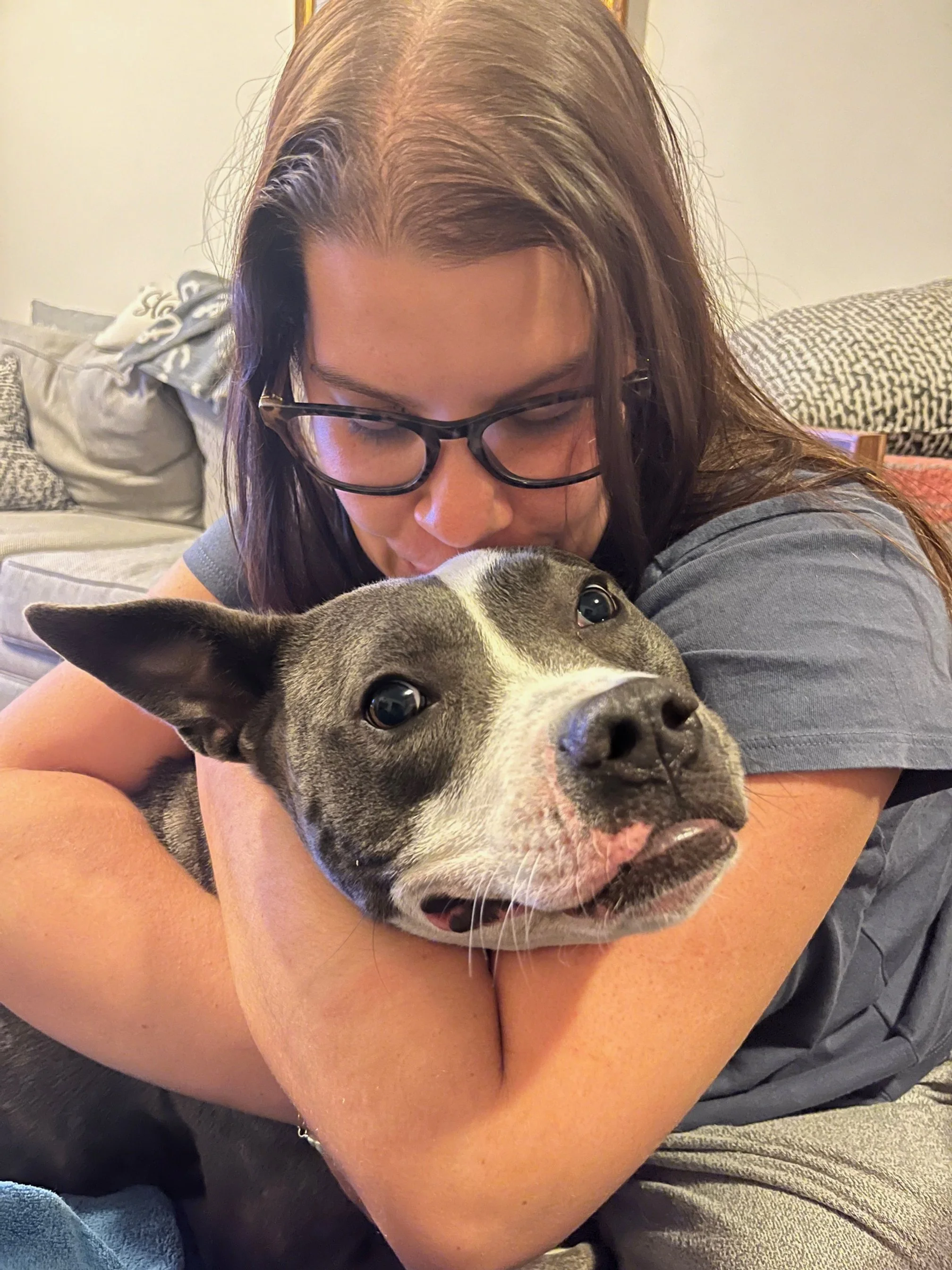 A woman with glasses hugging a gray and white dog.