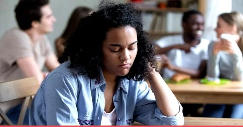 woman sitting slightly apart from group in social setting representing avoidance due to anxiety or fear of getting sick