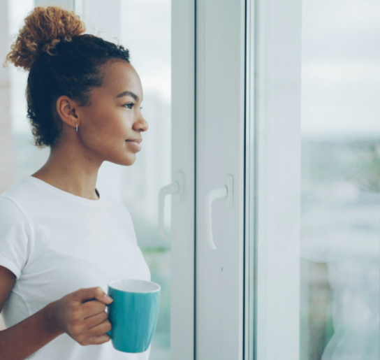 woman standing by window holding coffee in calm, reflective moment representing awareness of overthinking or anxiety patterns