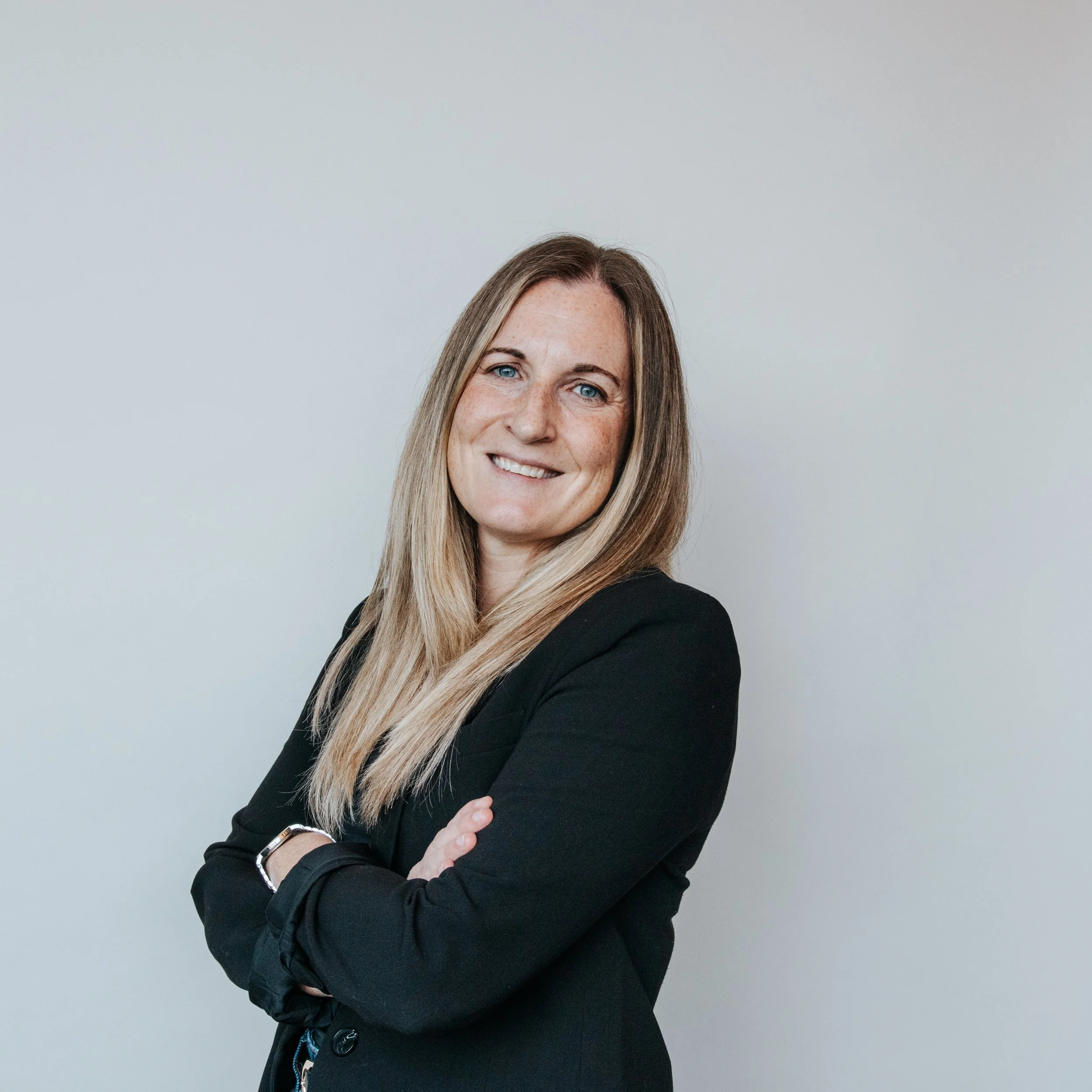 Professional woman with long blonde hair wearing a black blazer, smiling and crossing arms, posing against a plain light gray background.