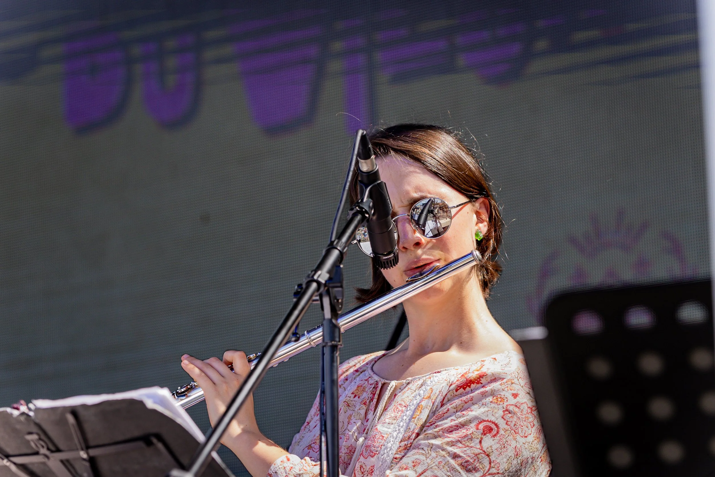 A woman wearing sunglasses and a patterned blouse playing a silver flute during a performance, with music stands and a large, blurred backdrop behind her.