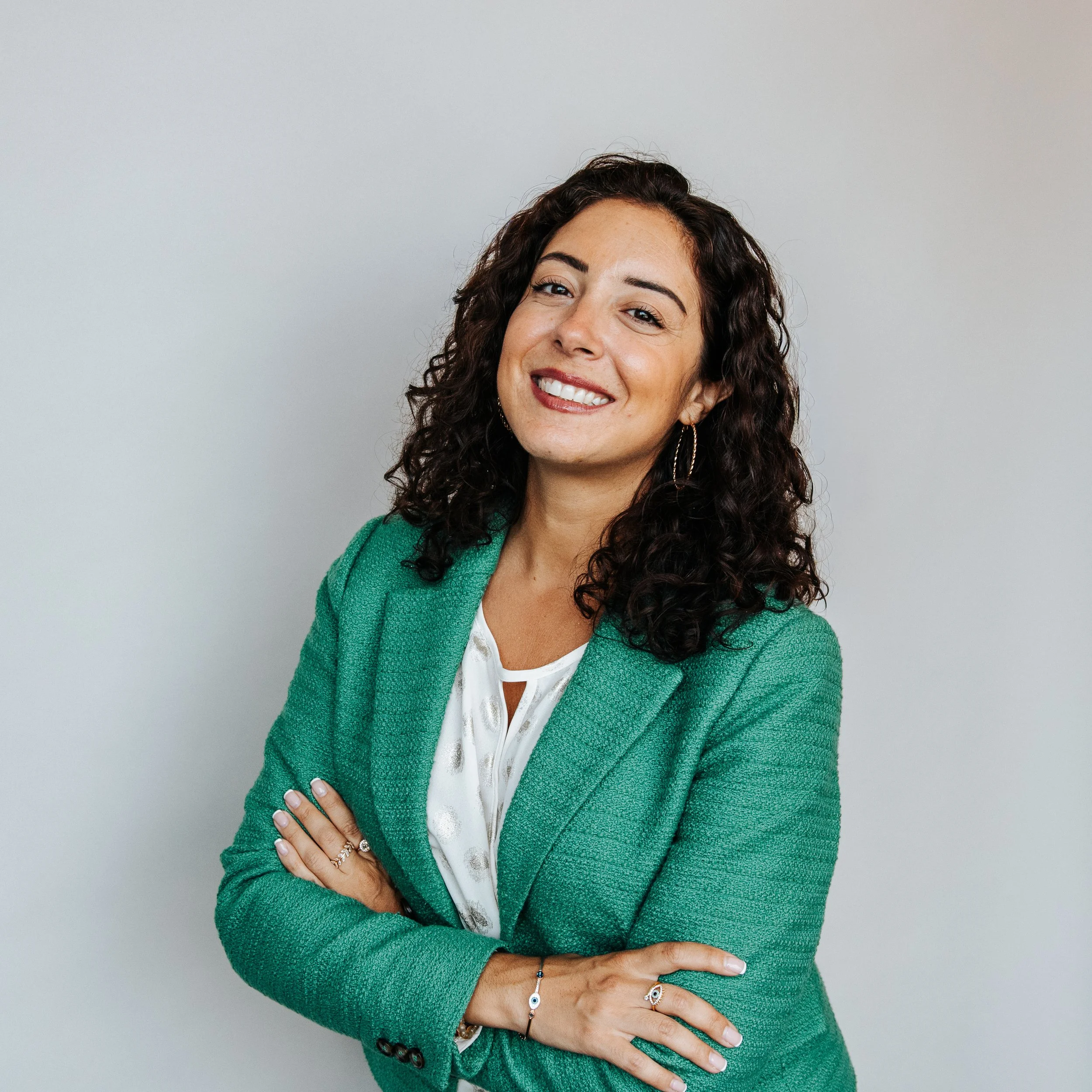 Portrait of a woman with curly dark hair, smiling, wearing a green blazer over a white top, standing against a plain gray wall.