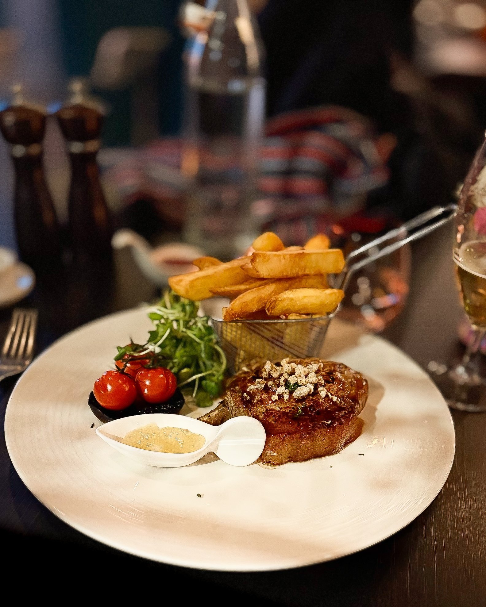 A plated meal with a grilled steak topped with herbs or spices, a small salad with cherry tomatoes, a portion of French fries in a small basket, a dollop of sauce in a spoon, and a glass of white wine.