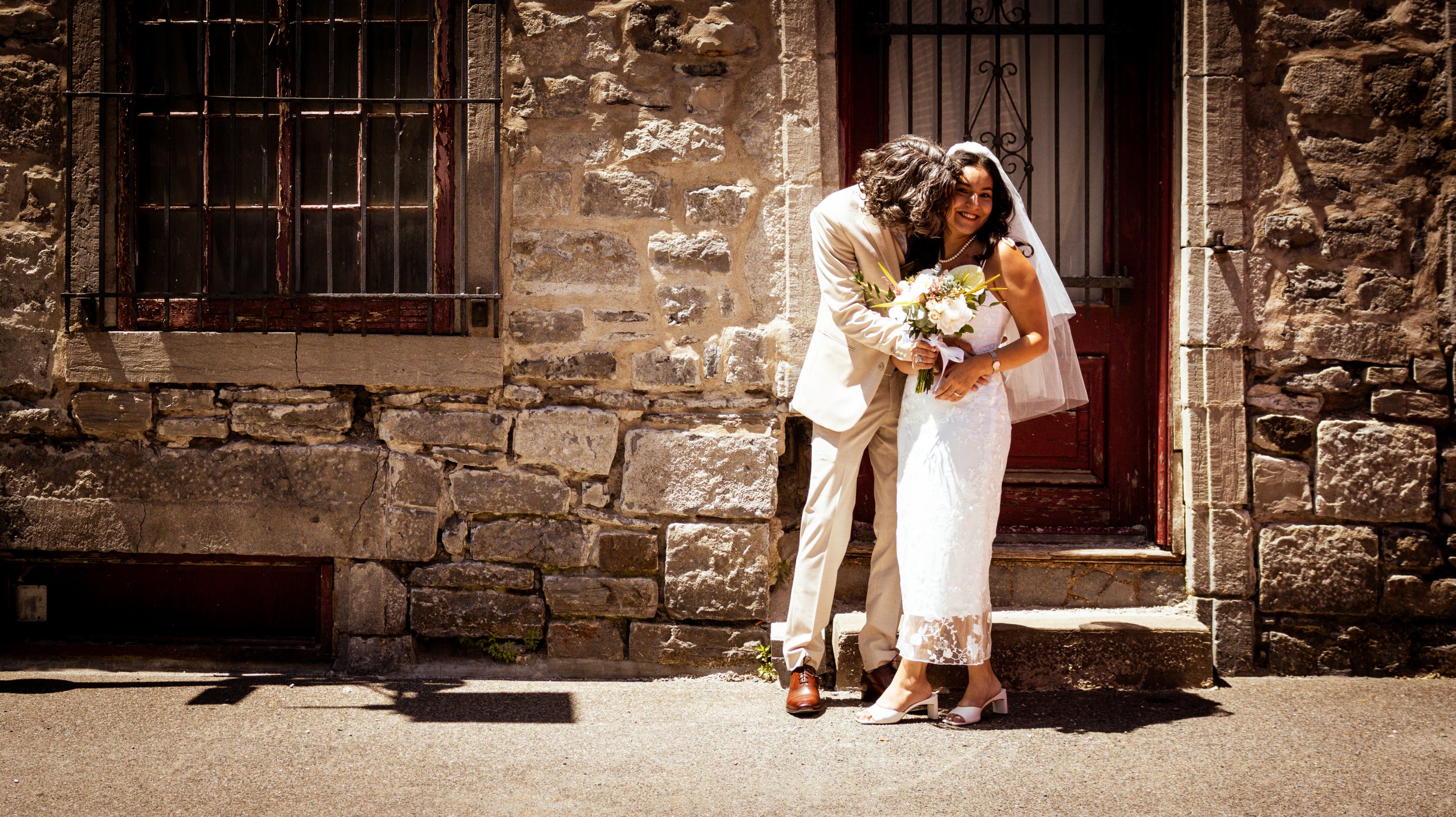 A bride and groom, dressed in wedding attire, stand close together outside a rustic stone building. The bride holds a bouquet of flowers and smiles, while the groom leans in towards her. Sunlight casts shadows on the ground.