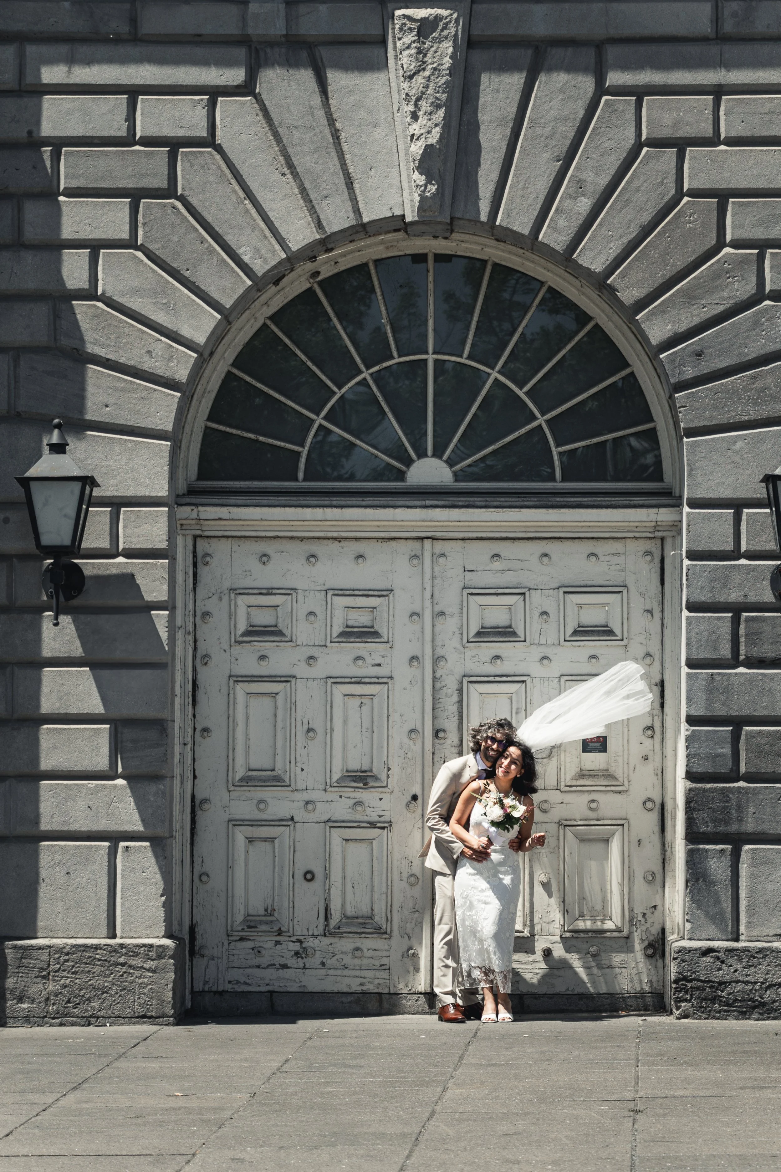 A couple in wedding attire standing in front of a large, weathered wooden door on a stone building, with a decorative arch and two wall-mounted lanterns on either side.