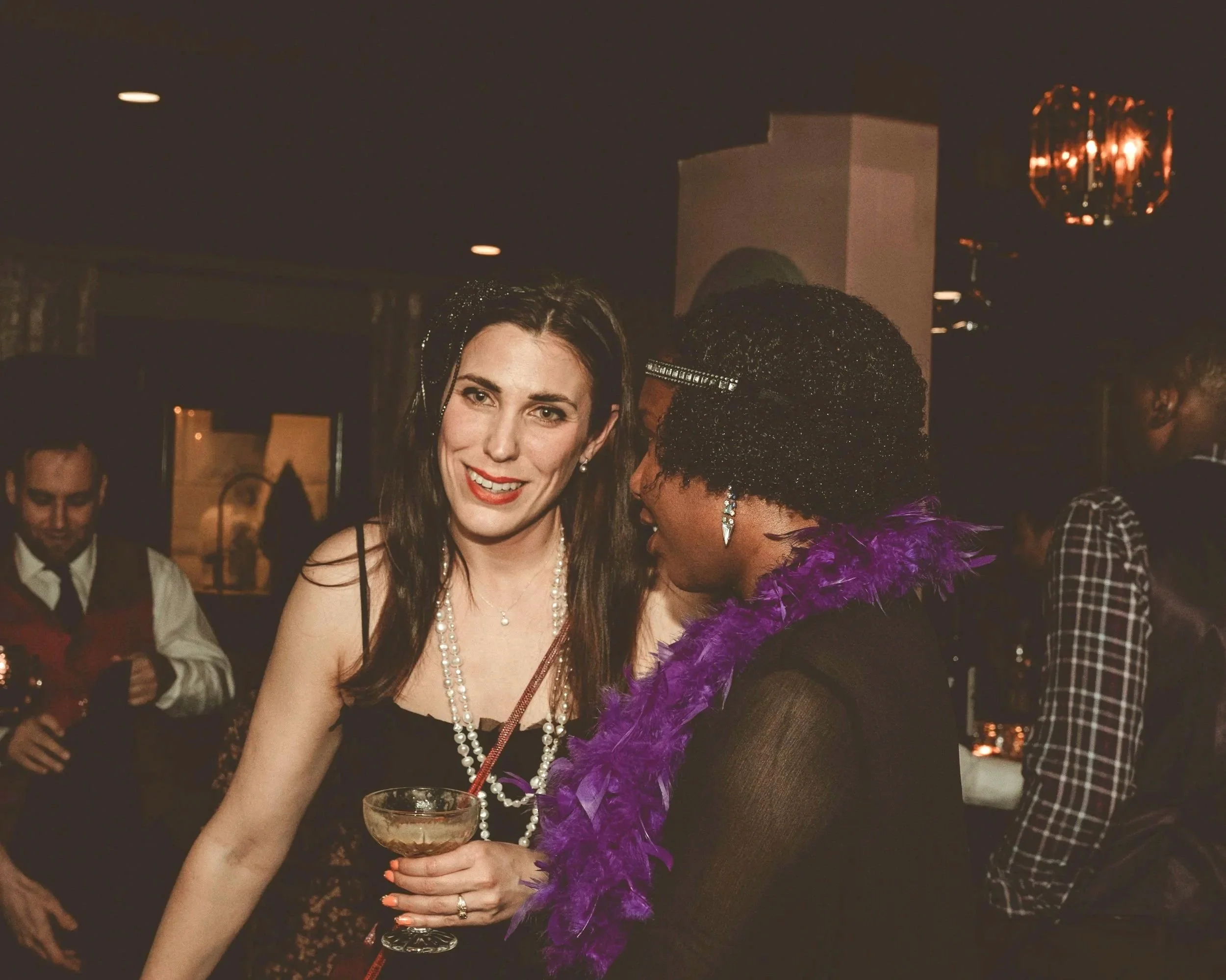 Two women dressed in party attire at a social event, one holding a cocktail, with other people in the background.