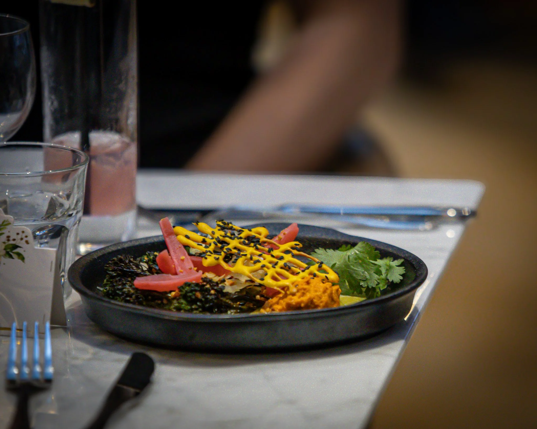 A black plate with a colorful summer roll garnished with yellow sauce and black sesame seeds, served with fresh cilantro and pickled ginger, on a white tablecloth at a table setting.