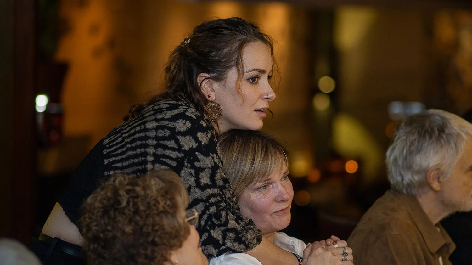 A woman with curly hair and earrings leans over a woman with hand clasped together in front of her, sitting at a table, in a warmly lit room with blurred background.