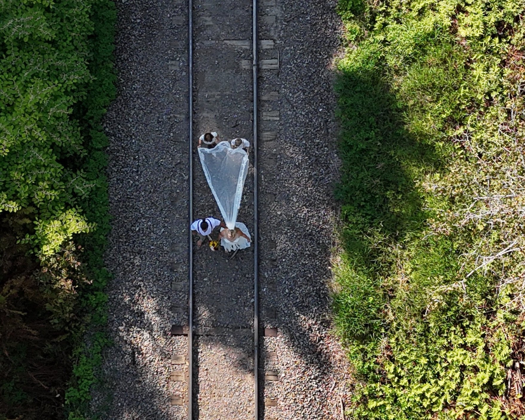 Aerial view of a couple wearing wedding attire walking along train tracks surrounded by green trees and bushes.
