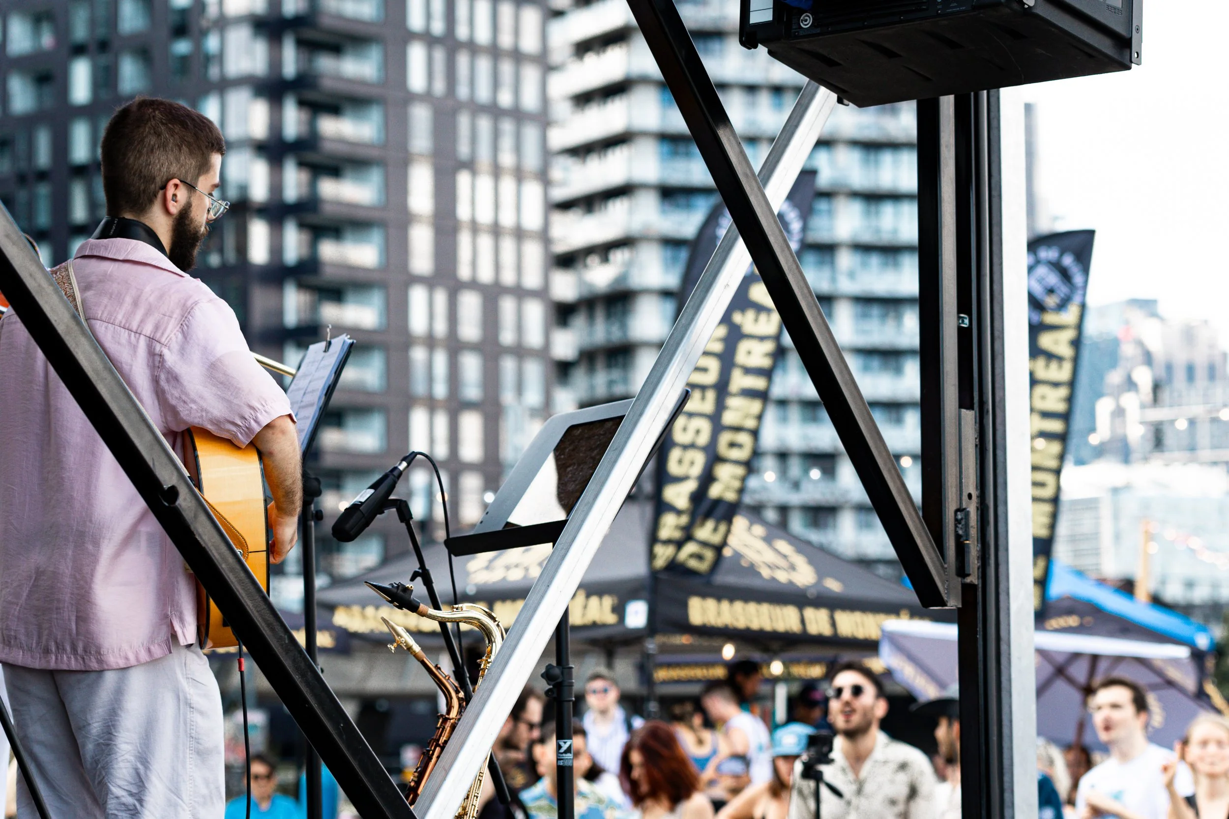 A man playing guitar and singing at an outdoor event with a crowd, city buildings, and banners in the background.
