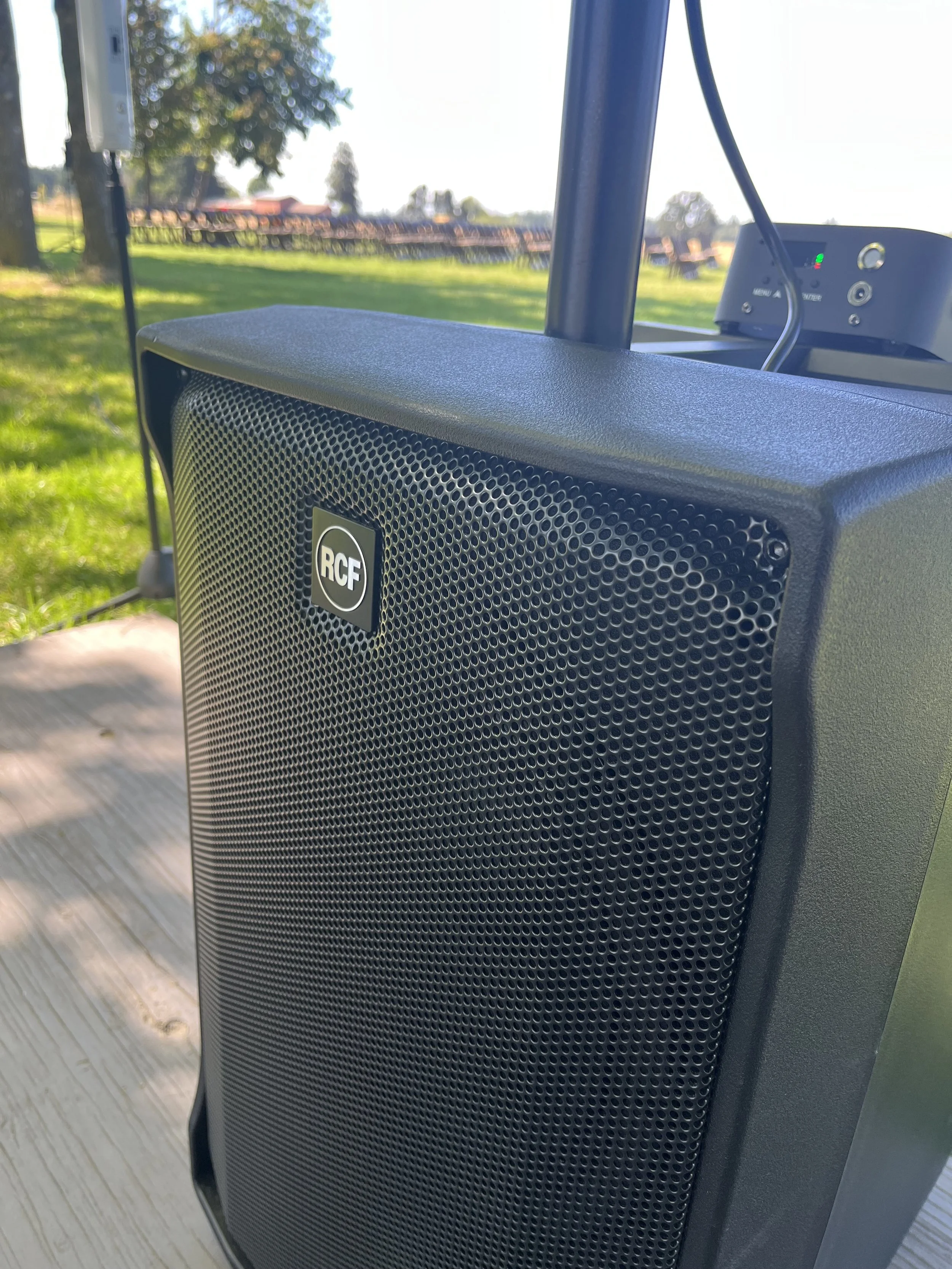 Close-up of a black RCF speaker outdoors on a wooden surface, with a grassy field, trees, and a seating area in the background.