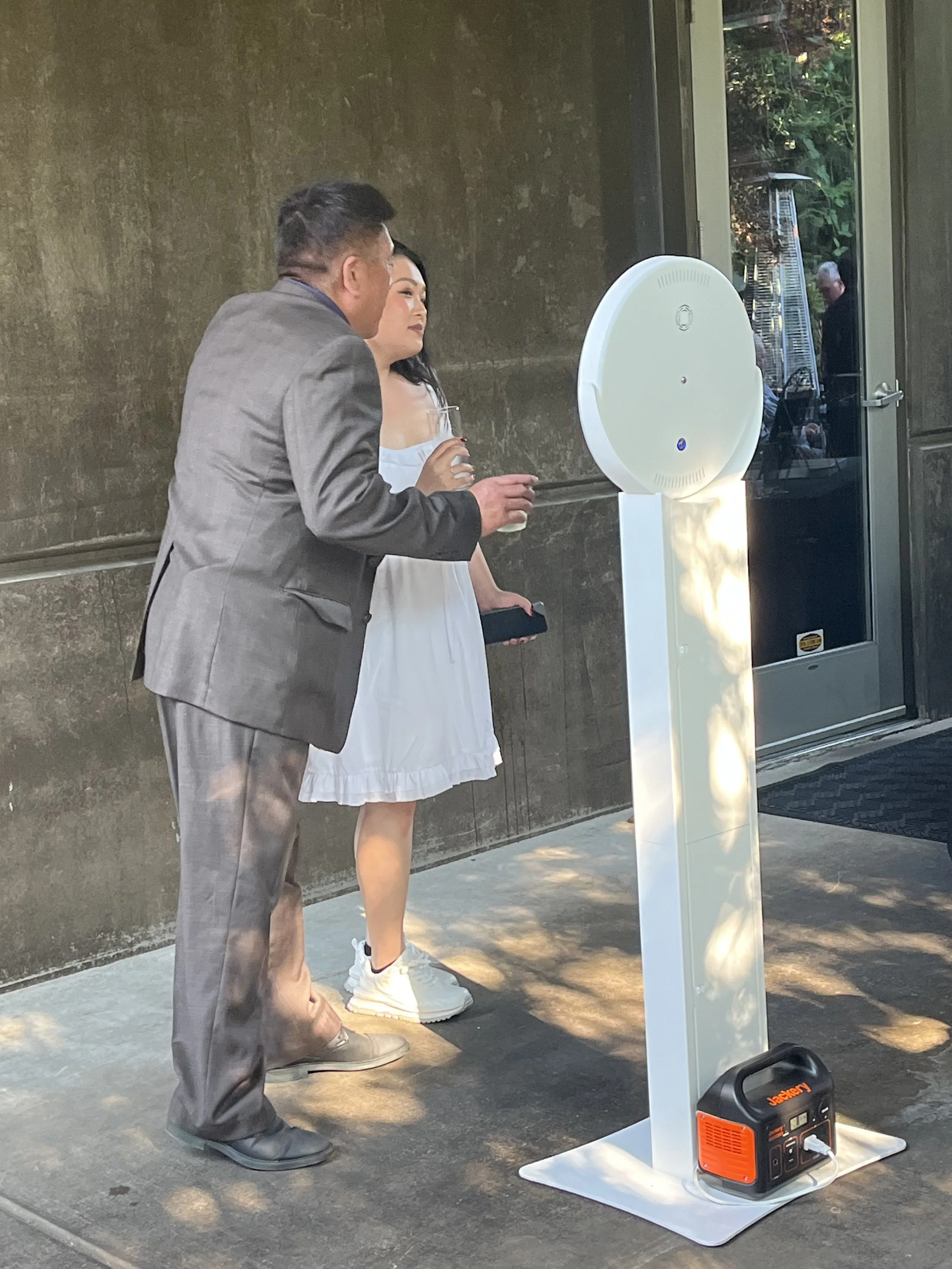 A man and woman stand outdoors next to an air purifier on a sidewalk, with a glass door in the background reflecting trees and people.