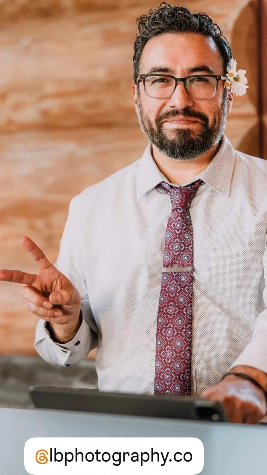 A man with glasses and a beard, wearing a white shirt and a patterned red tie, is smiling and making a peace sign with his right hand. He has a small white flower tucked behind his right ear. The background features a wooden wall. There is a watermar