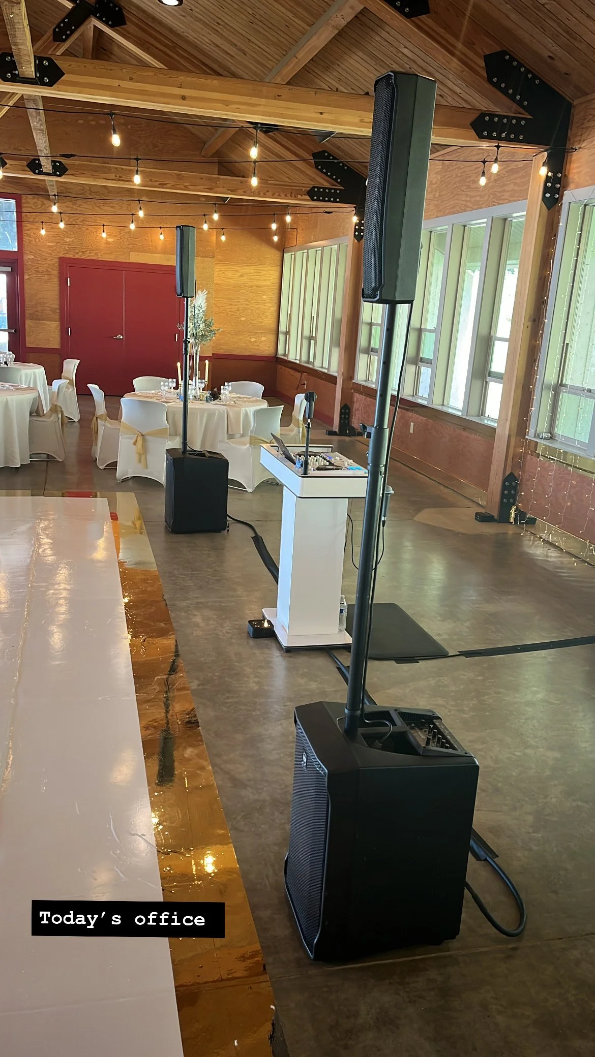 An event space with round tables covered in white tablecloths, decorated with candles and floral centerpieces. The space has a wooden ceiling with string lights, large windows, and a polished floor. There are two large standing speakers, a DJ setup,