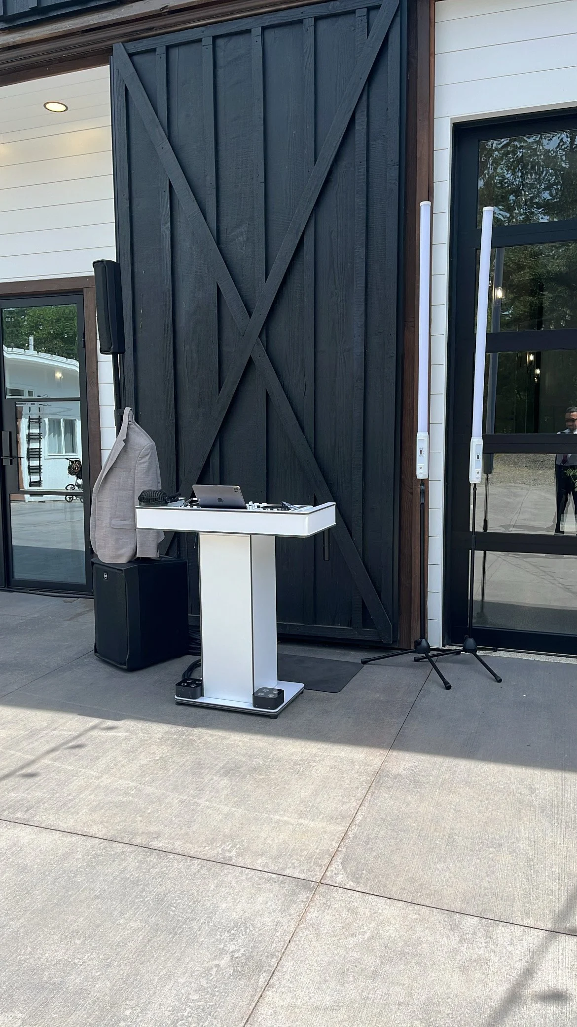 An outdoor DJ setup with a small white table holding DJ equipment, a speaker, a beige bag, two microphone stands, and a person’s reflection visible in the glass door.