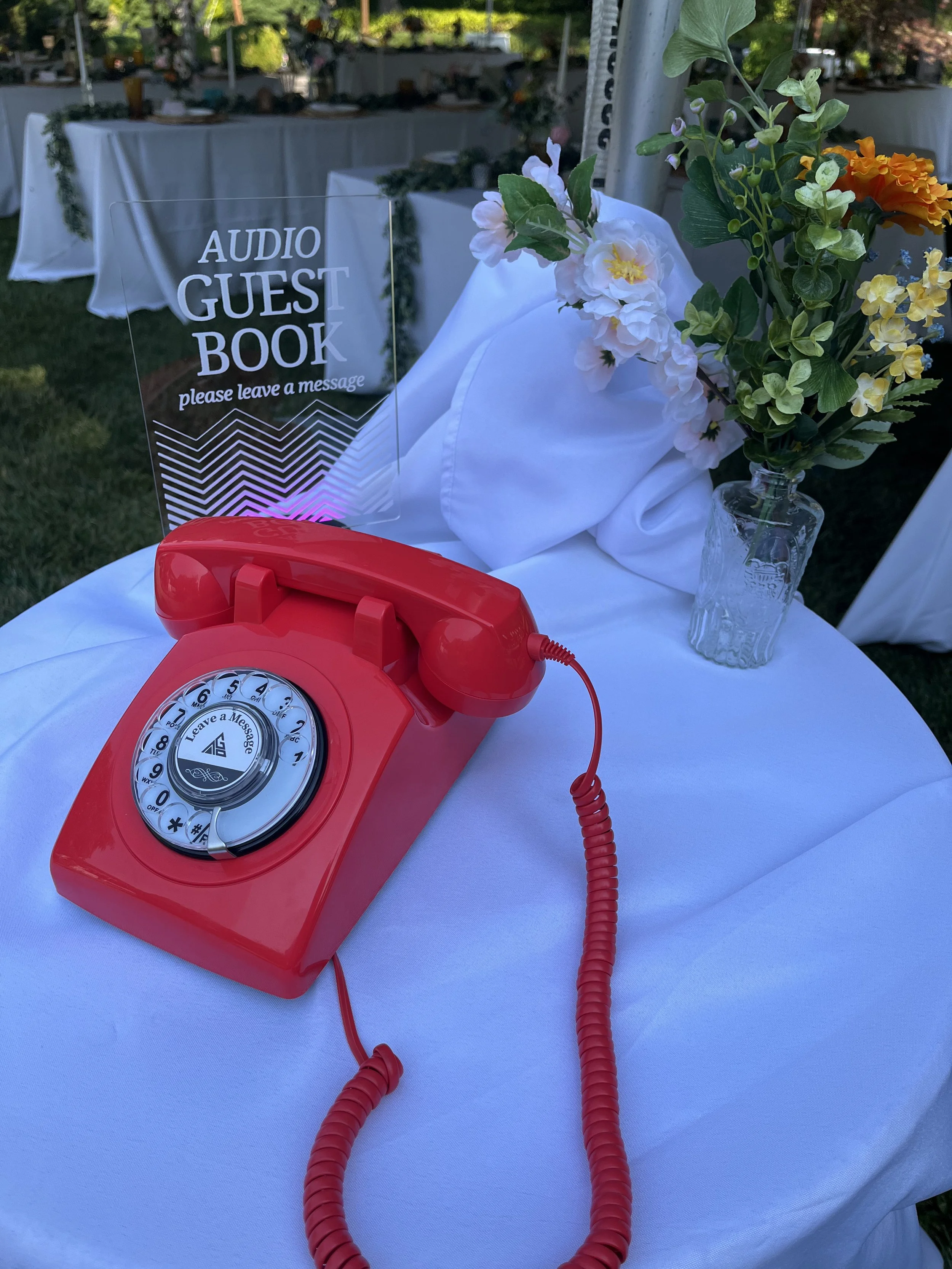A vintage red rotary phone is placed on a white table with a sign that reads 'Audio Guest Book, please leave a message.' next to a vase of mixed flowers, including white, yellow, and orange blooms.