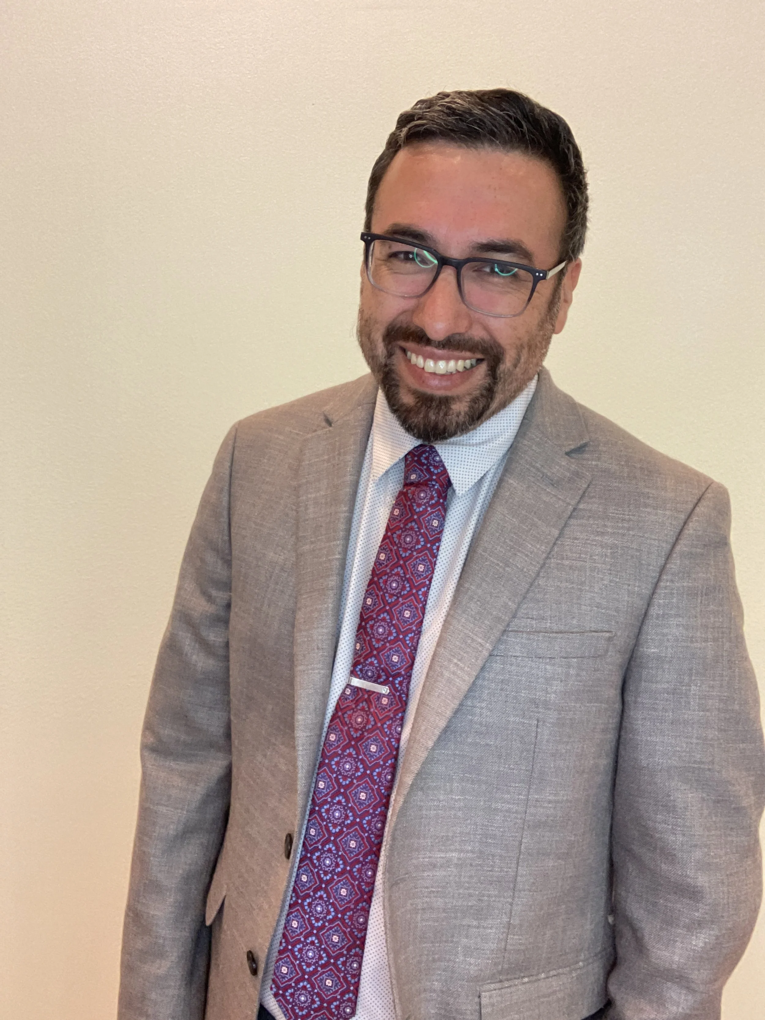 A man with glasses, a beard, and dark hair smiling, dressed in a light brown suit, white shirt, and patterned red tie, standing against a plain light-colored wall.