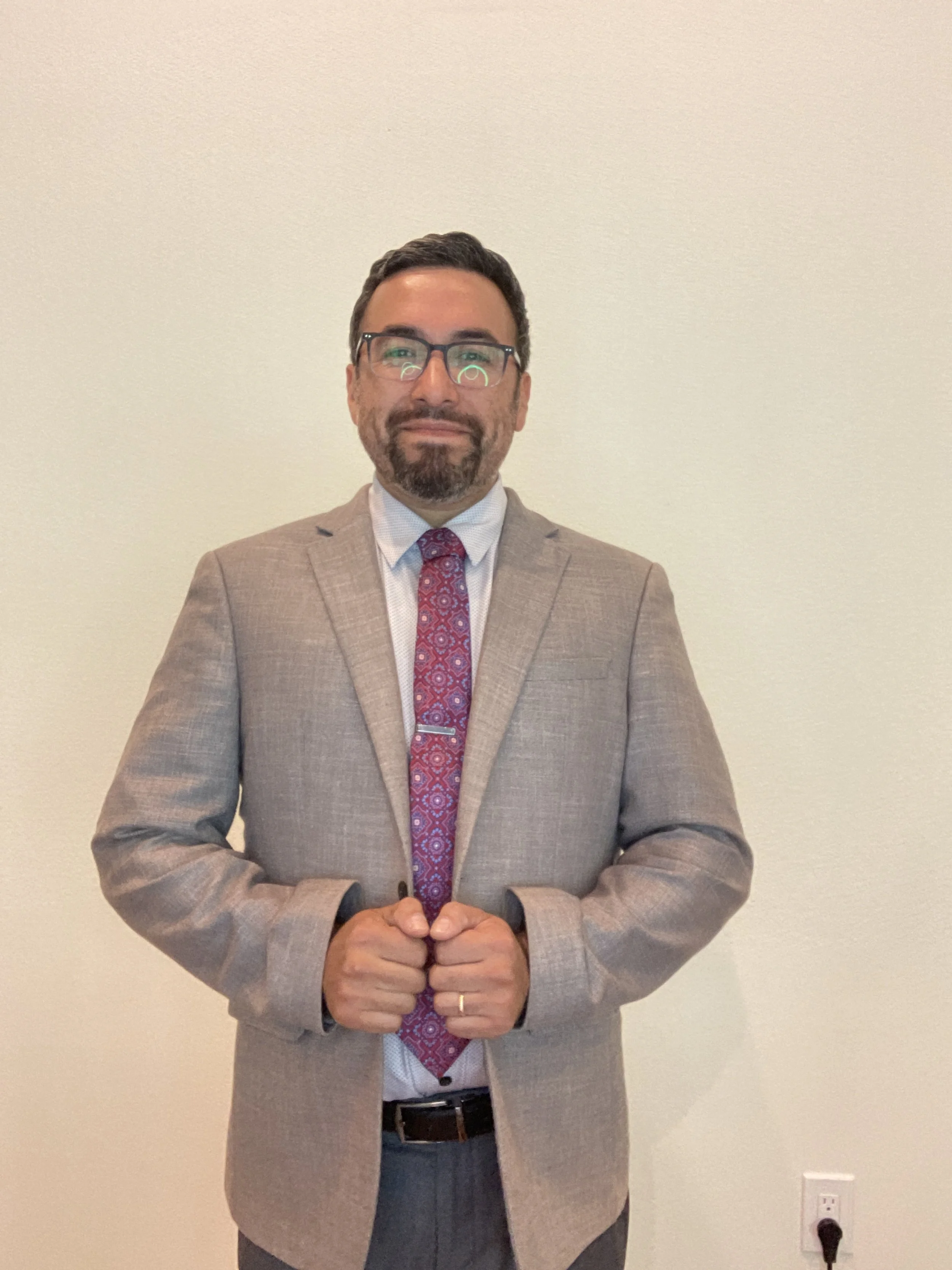 A man in a gray suit, white shirt, and patterned red tie standing against a plain light-colored wall.