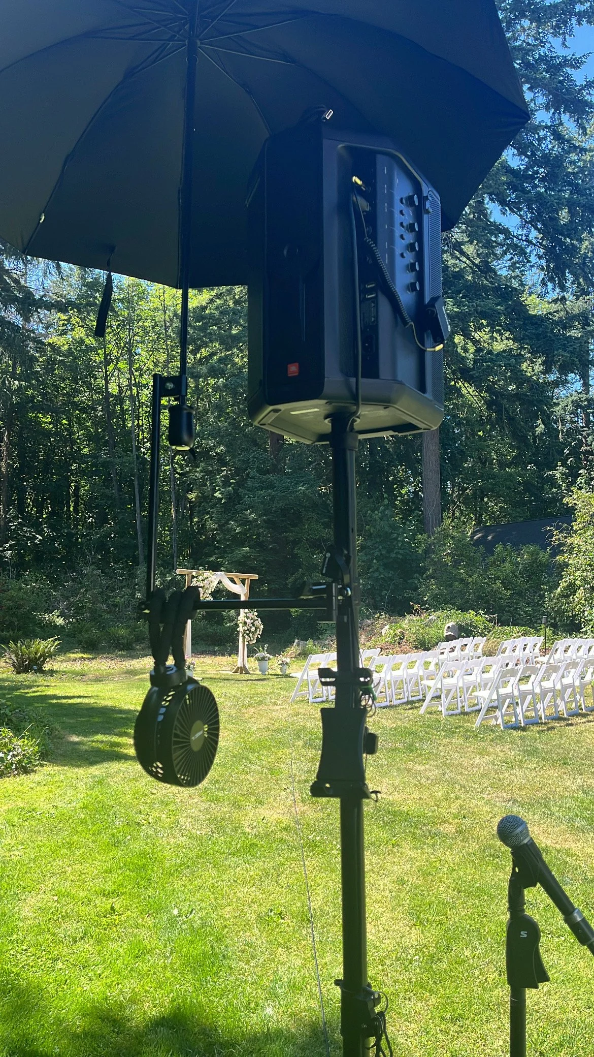 Outdoor wedding setup with white chairs arranged on a green lawn, floral arch, and a speaker mounted on a stand under a black umbrella, with a microphone on a stand nearby.