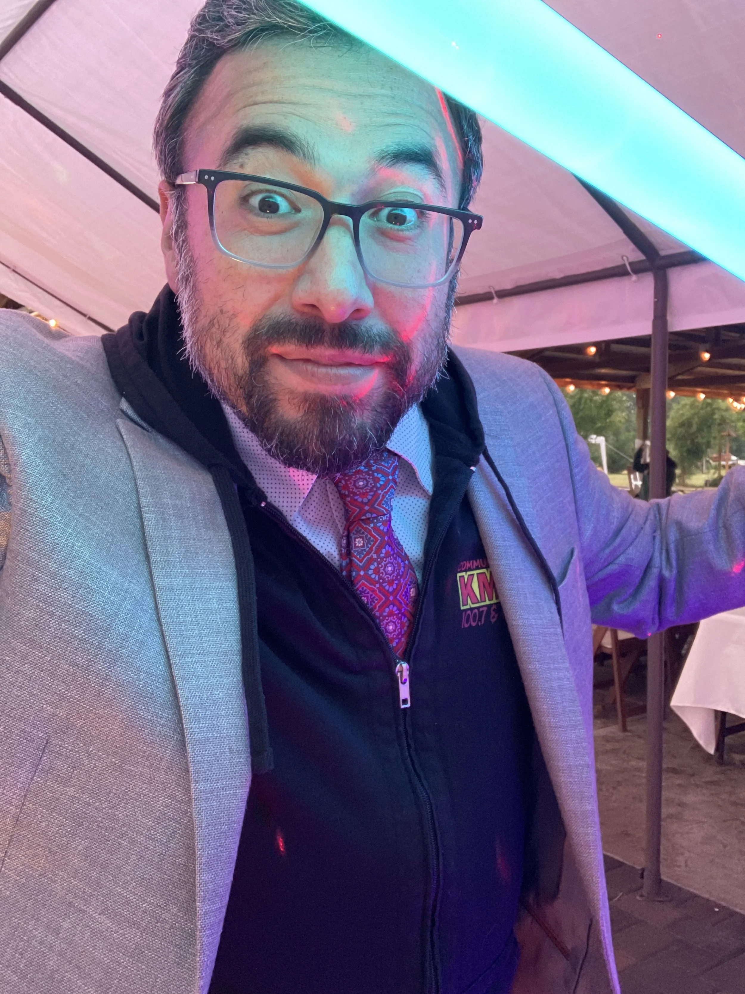 A man with glasses, a beard, and mustache taking a selfie at an outdoor event, wearing a blazer over a black hoodie with a logo, and a patterned tie under lighting that creates a colorful glow.