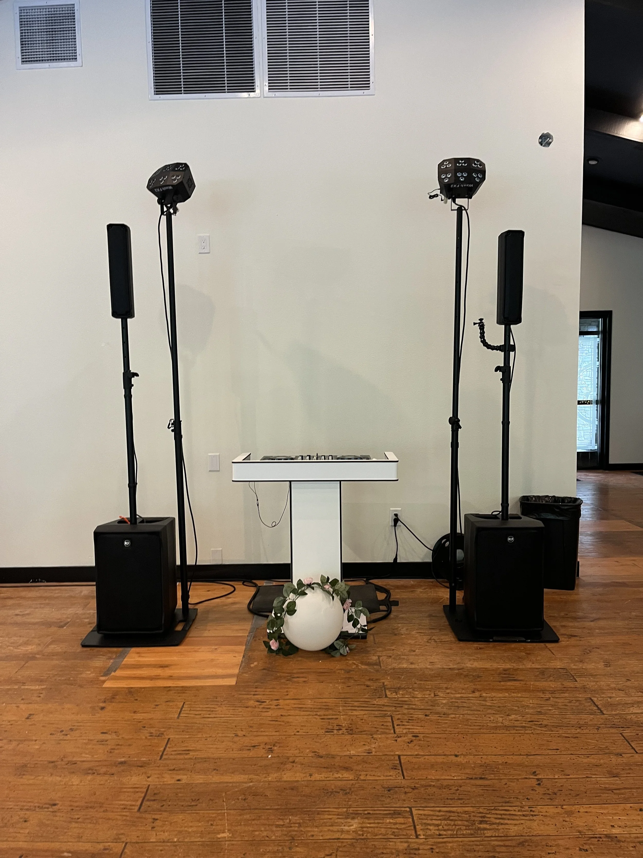Wedding DJ setup with speakers, lights, and a keyboard on a stand, decorated with a flower-adorned vase, on hardwood floor.
