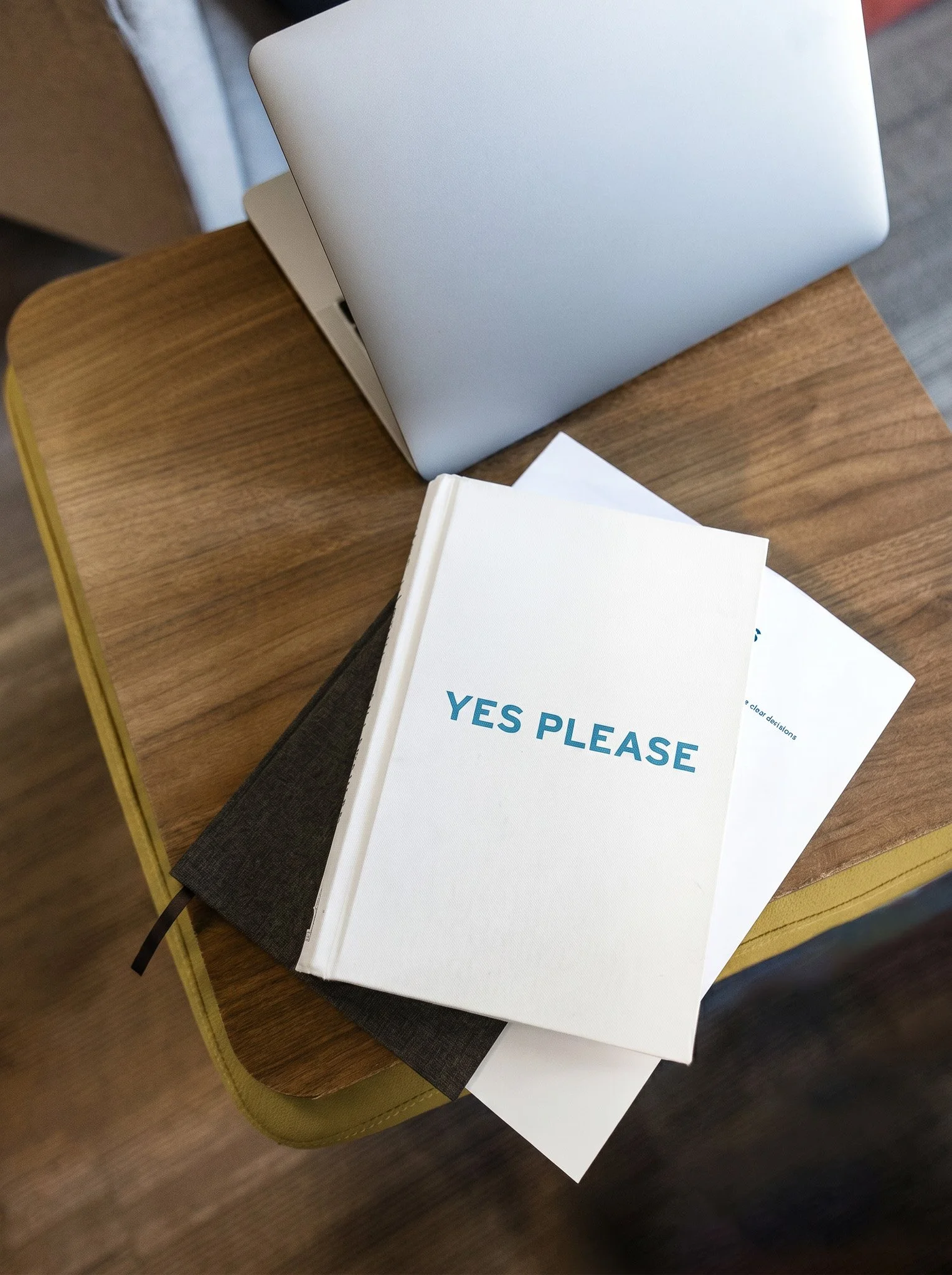 A silver laptop and the book, "Yes Please" are on a square wood table.