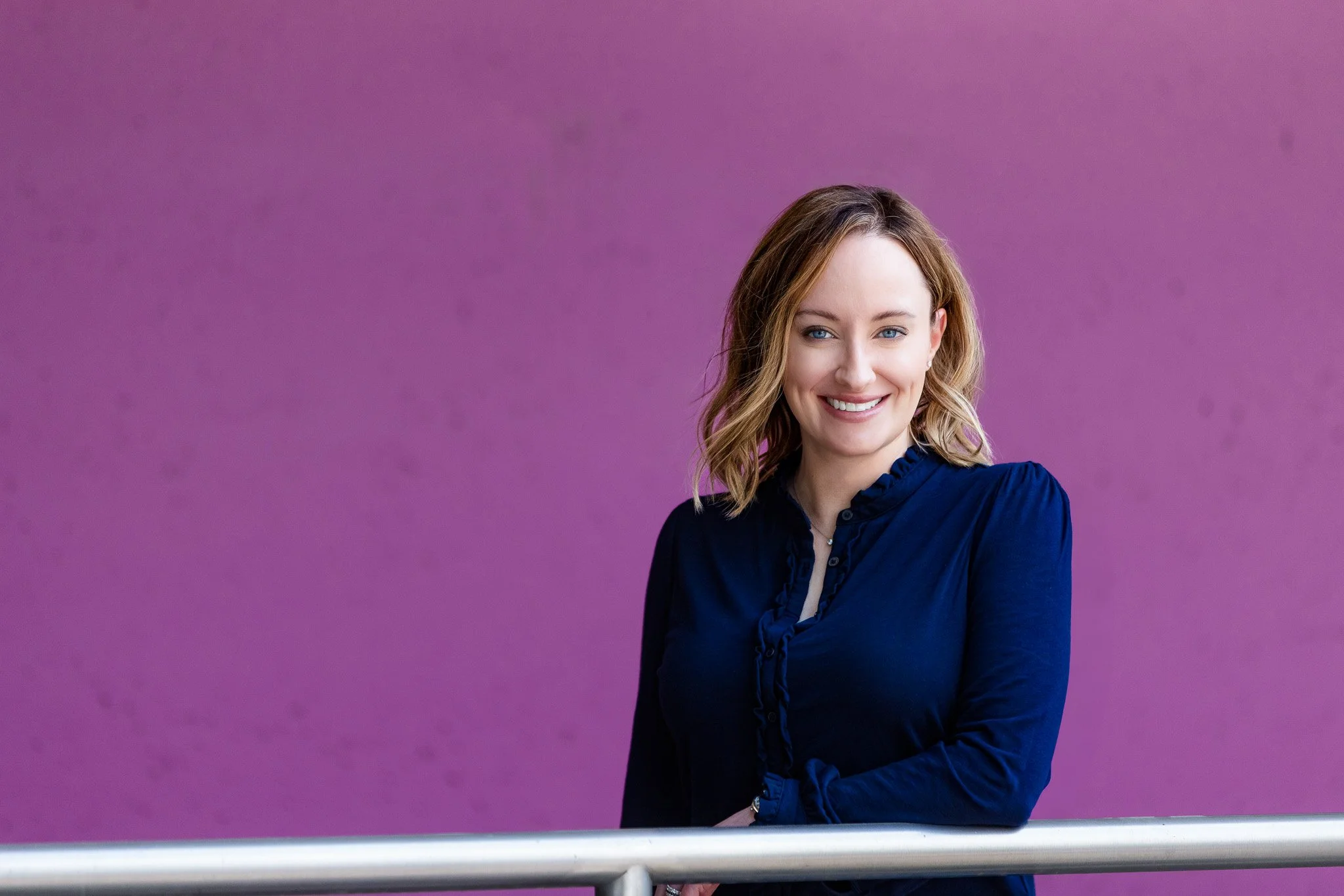Melissa's looking at the camera and wearing a navy shirt standing in front of a purple painted concrete wall.
