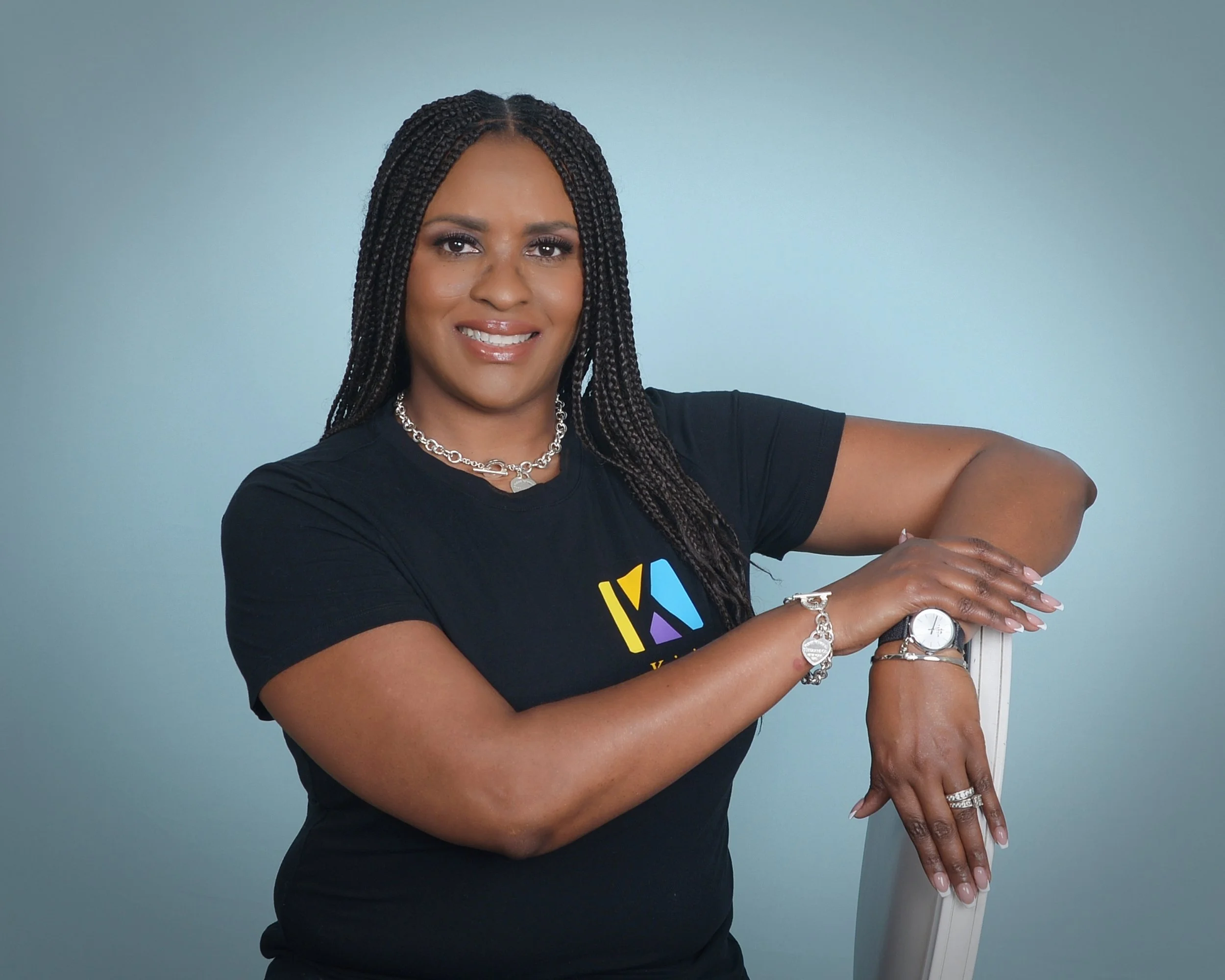 A woman with braided hair wearing a black t-shirt, jewelry, and a watch, leaning on a white chair against a plain light blue background.