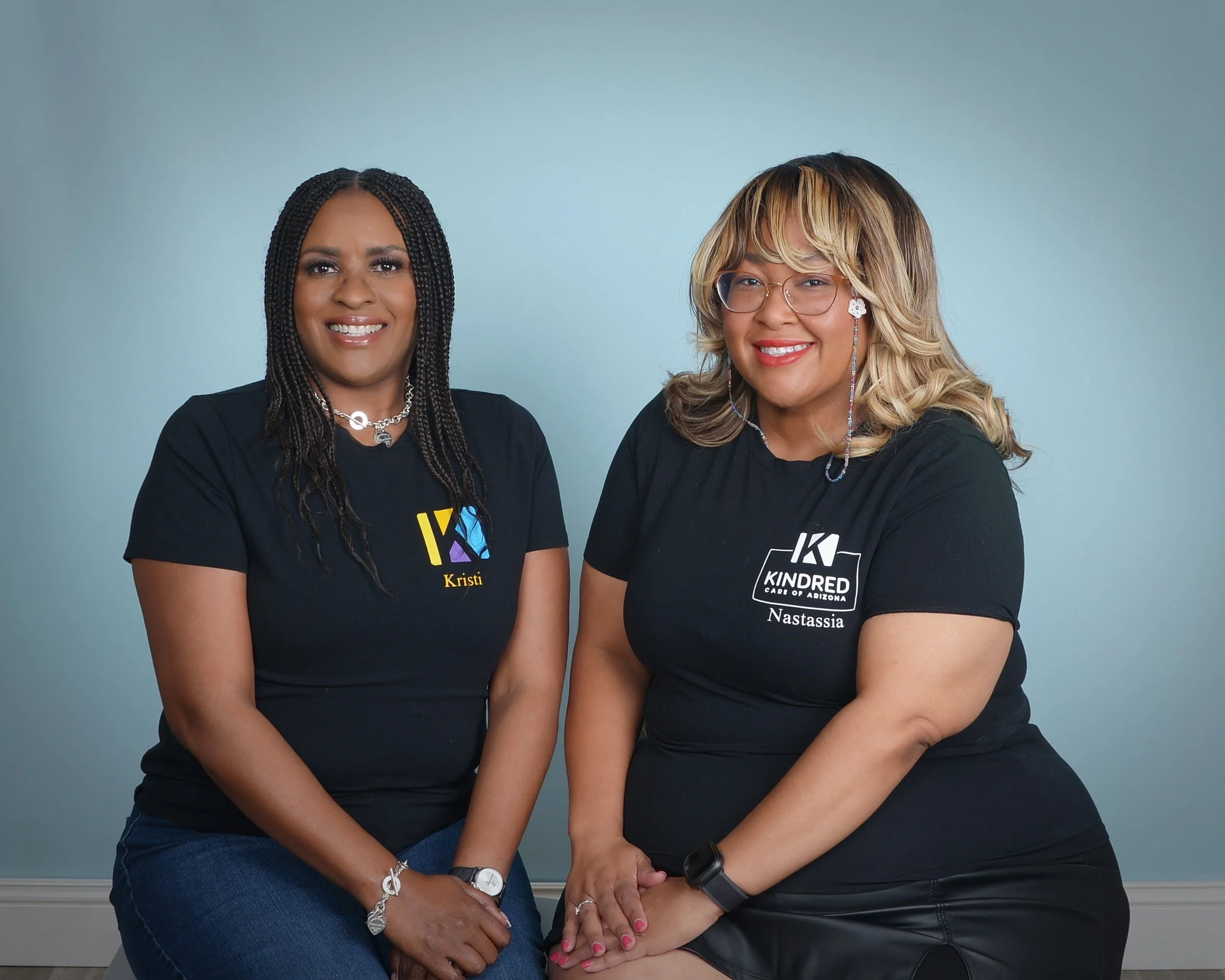 Two women sitting side by side in front of a plain wall, both wearing black shirts with logos and their names.