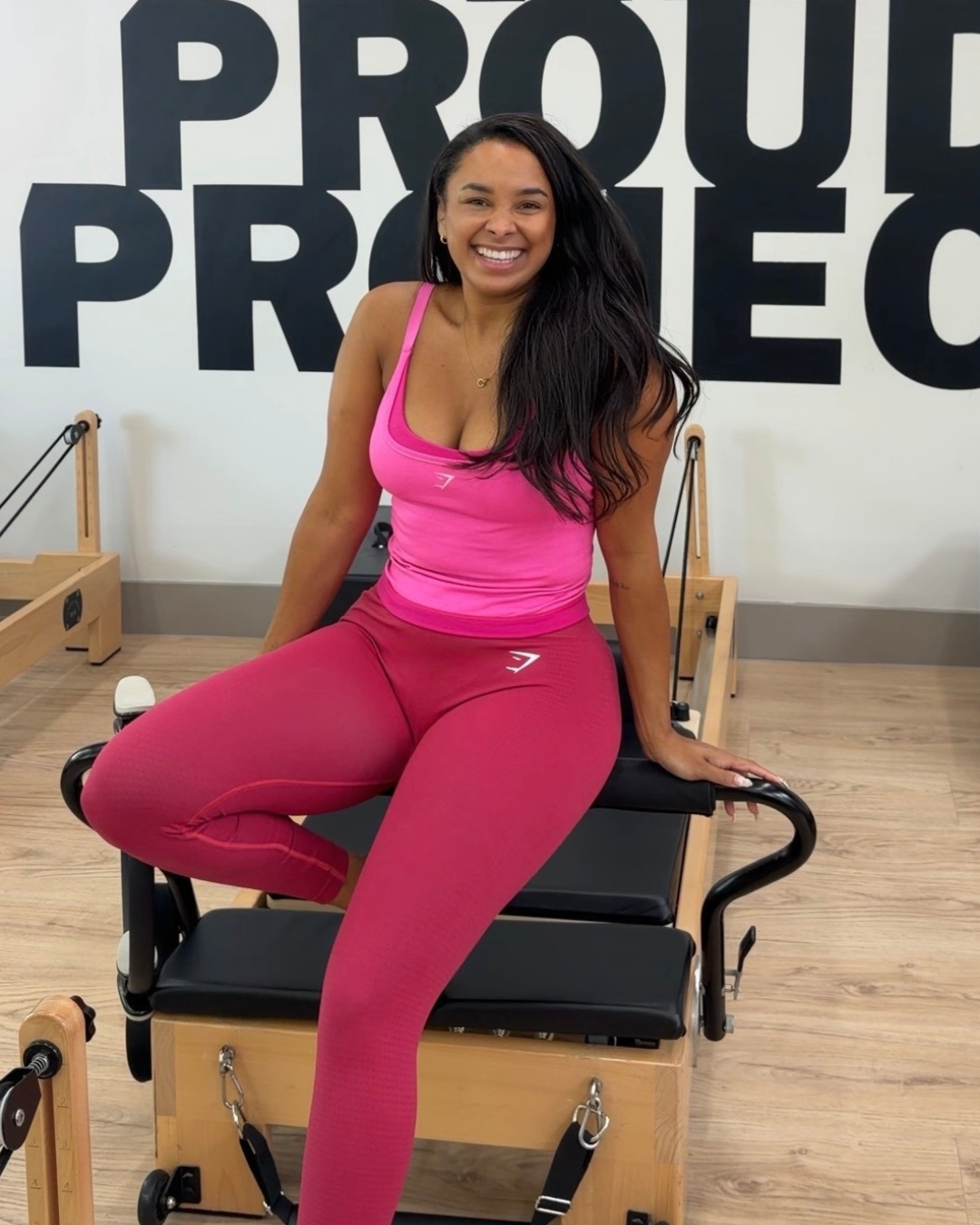 Woman sitting on Pilates reformer machine, smiling, wearing pink workout clothes, in a fitness studio with a wall reading 'PROUD PRO'.