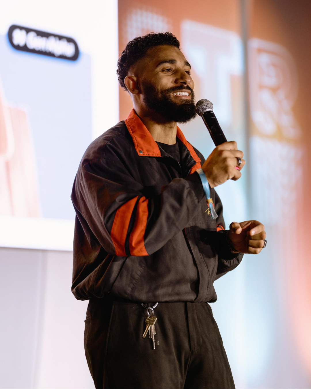 A man with a beard and curly hair holding a microphone and smiling while giving a presentation in front of a large screen.
