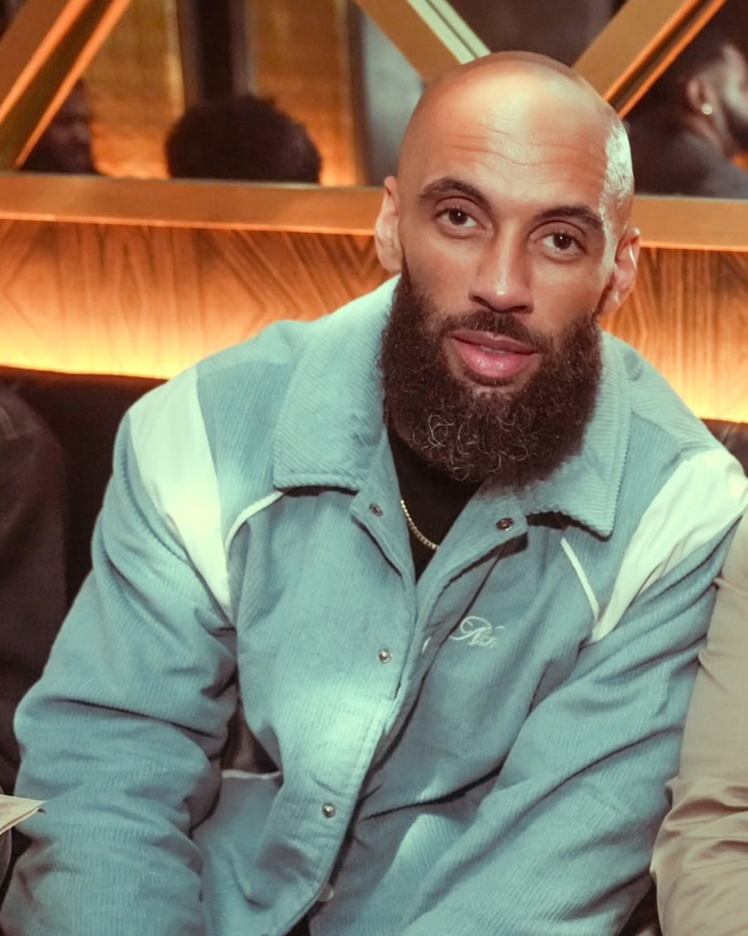 A man with a bald head and full beard, wearing a light-colored jacket, sitting indoors in a restaurant or cafe.