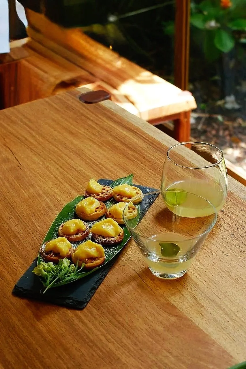 A wooden table with a black slate serving platter holding eight small appetizers topped with yellow sauce, garnished with a sprig of herbs. Two glasses of greenish-yellow liquid sit beside the platter. In the background, a wooden bench and greenery are visible.