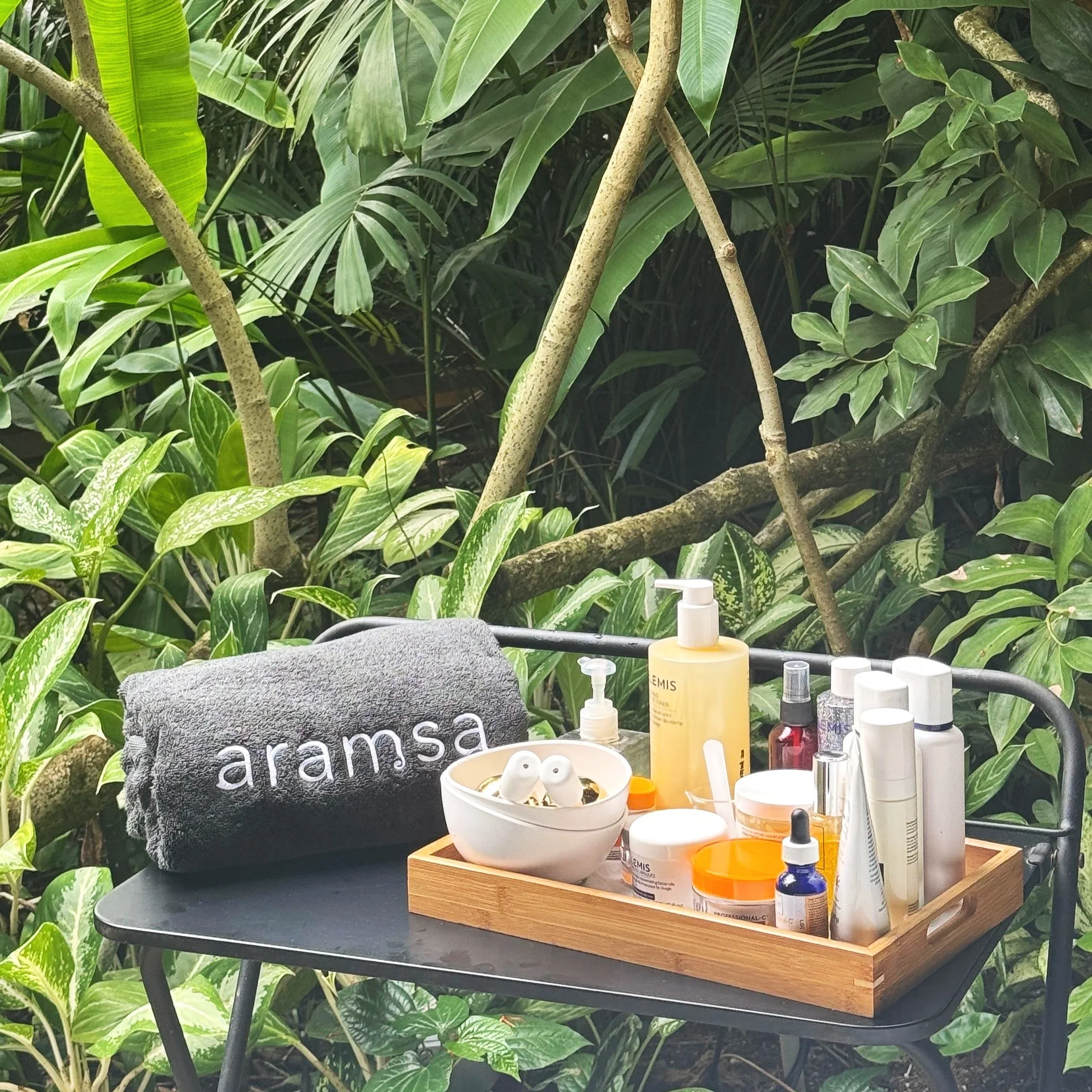 A black table outdoors with a gray towel embroidered with 'aramsa', a wooden tray containing skincare and beauty products, a white bowl with earbuds, and a small white spray bottle, surrounded by lush green plants.