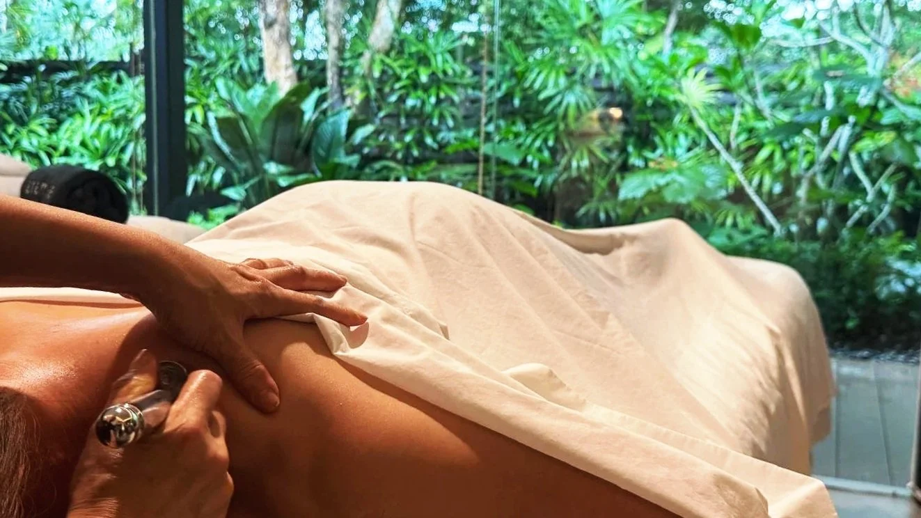 A person is receiving a massage from a massage therapist in a room with large windows and lush green plants outside.