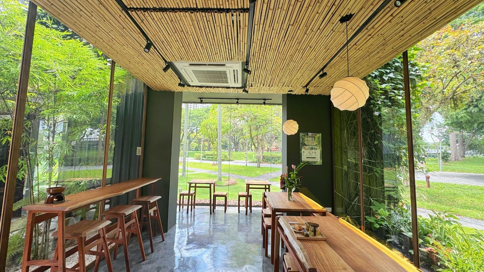 Interior of a modern cafe with large glass windows showing green trees and grass outside. Wooden tables and chairs are arranged inside, with some small hanging pendant lights and an air conditioning unit on the ceiling.