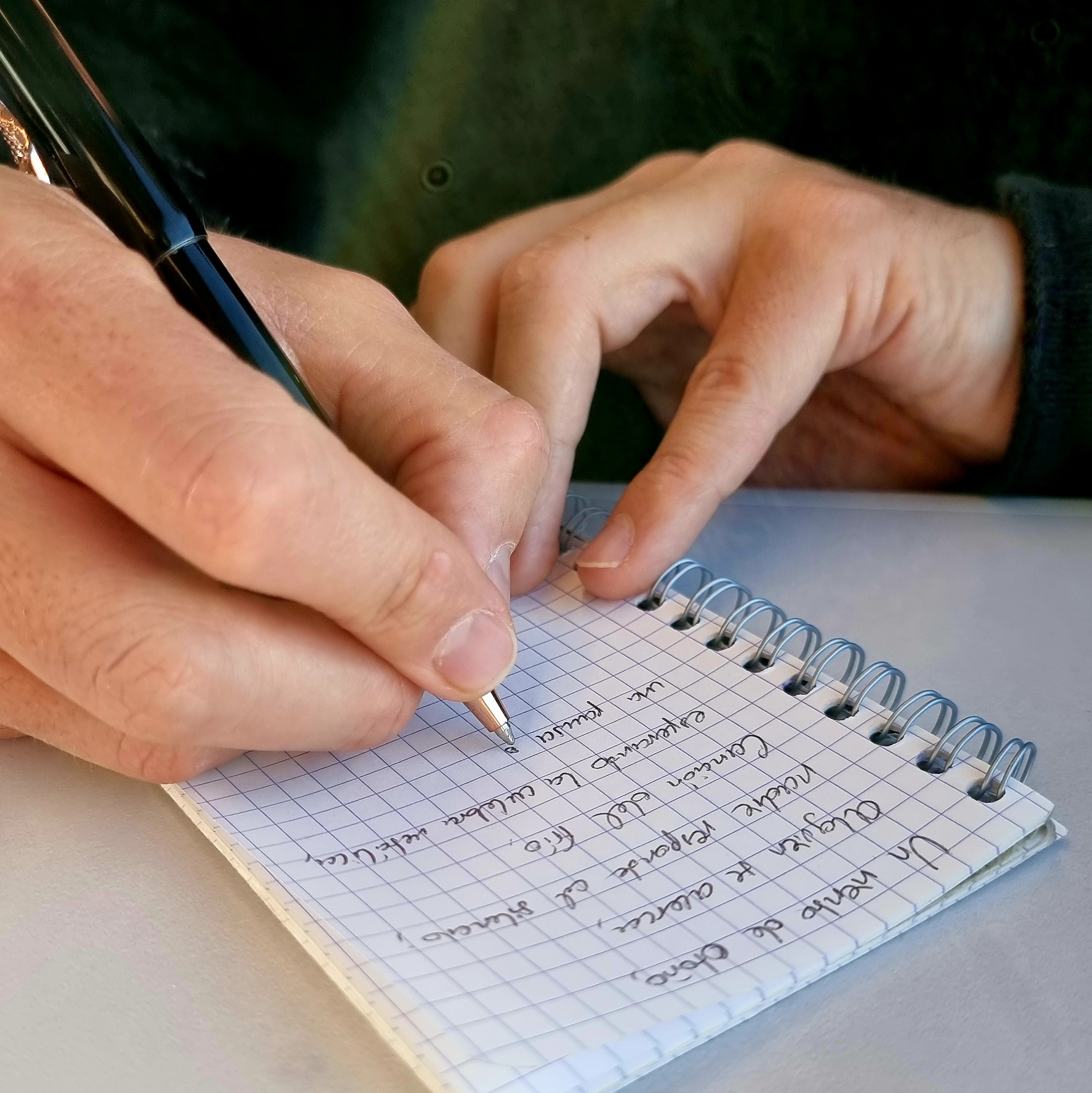A person writing in a spiral notebook with a black pen, the notebook has blue grid paper and the text appears handwritten.