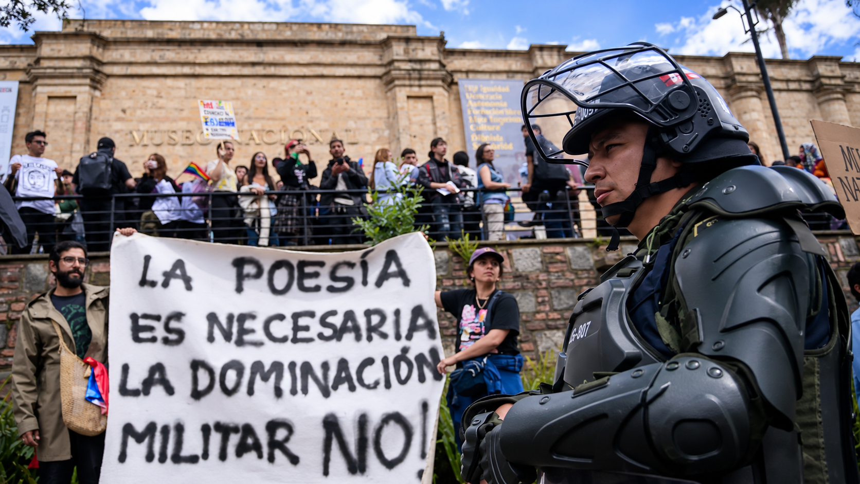 A riot police officer faces protesters holding a banner reading “Poetry is necessary, military domination is not,” symbolizing Colombia’s cultural war and ideological purge debate.