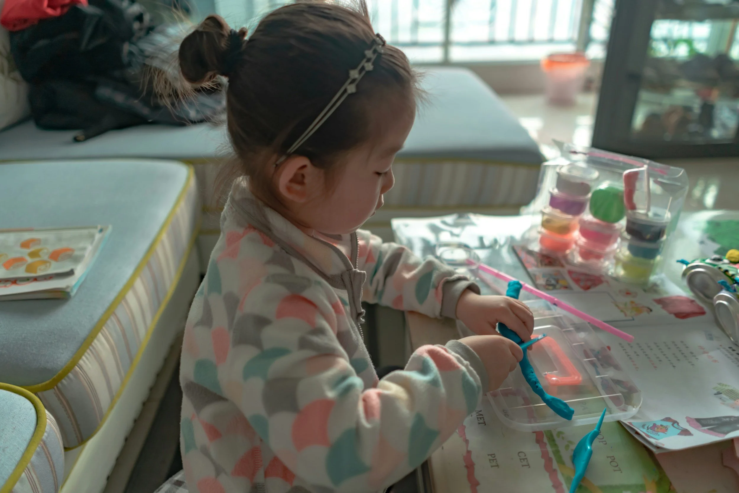 A young girl with a pastel polka dot hoodie sitting at a table, engaged in a craft activity with various colored paints and tools, in a room with large windows.
