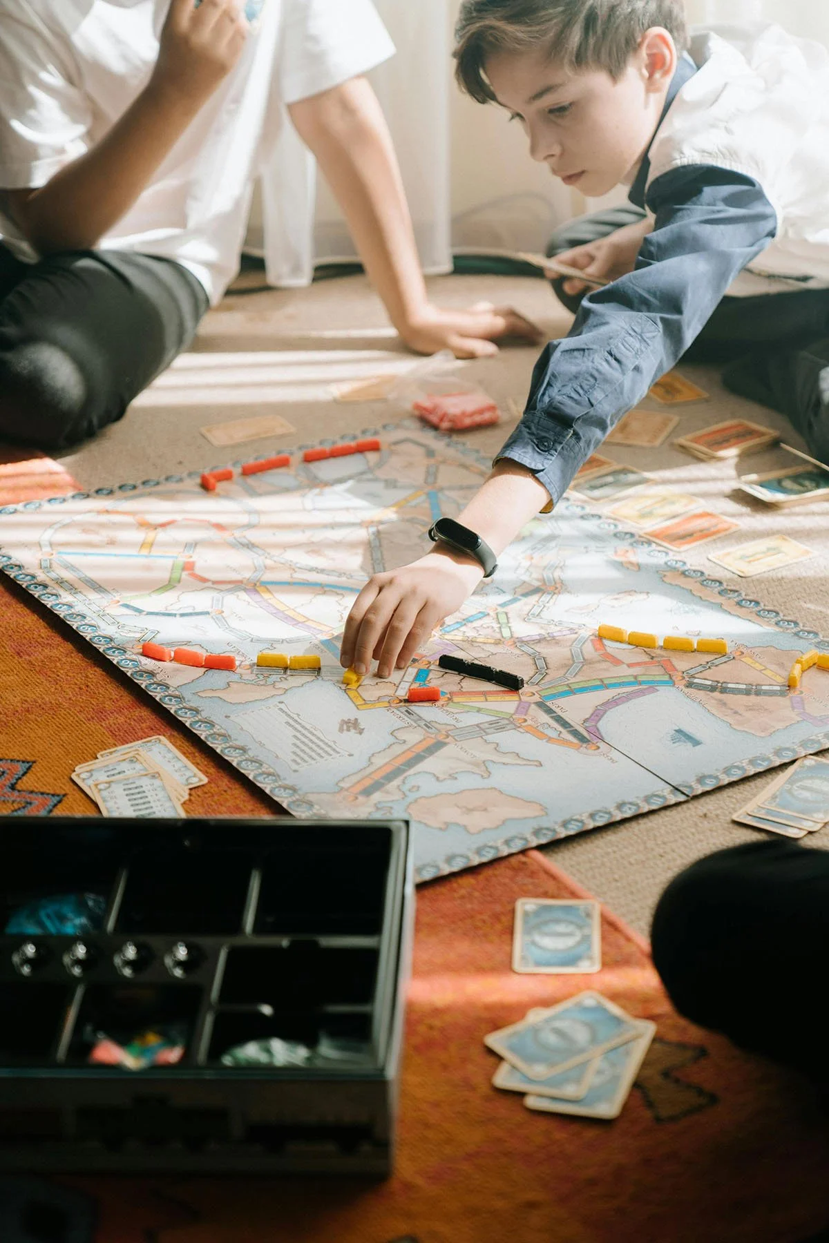 Children playing a board game on the floor, featuring a map with colored train pieces and cards.