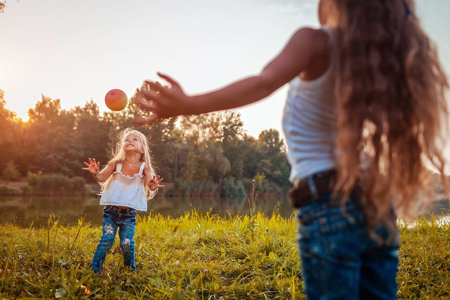 Two children playing catch with a ball by a lake, surrounded by greenery and trees, during sunset.