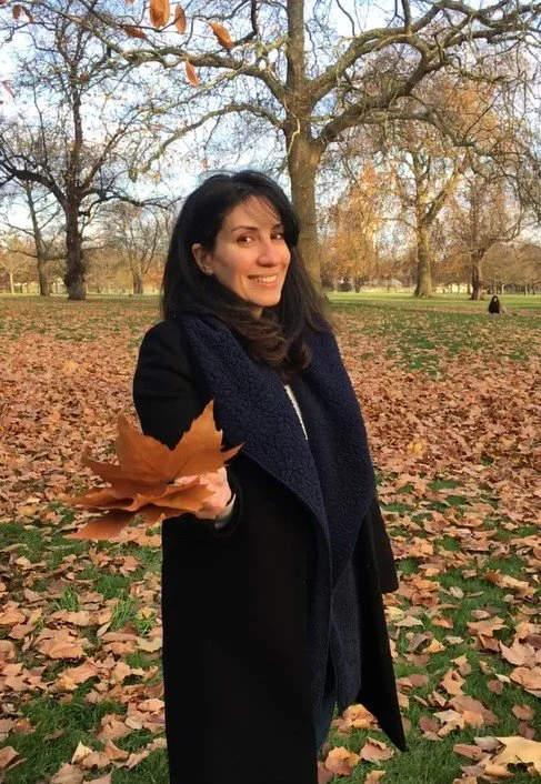 A woman in a black coat holding fall leaves in a park with trees and fallen leaves on the ground.