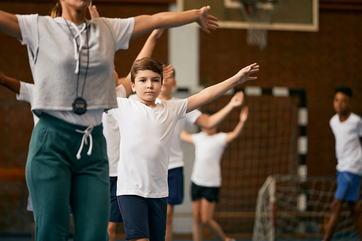 Children and instructor performing exercises in a gym class, wearing athletic clothing, focus on a young boy in a white shirt.